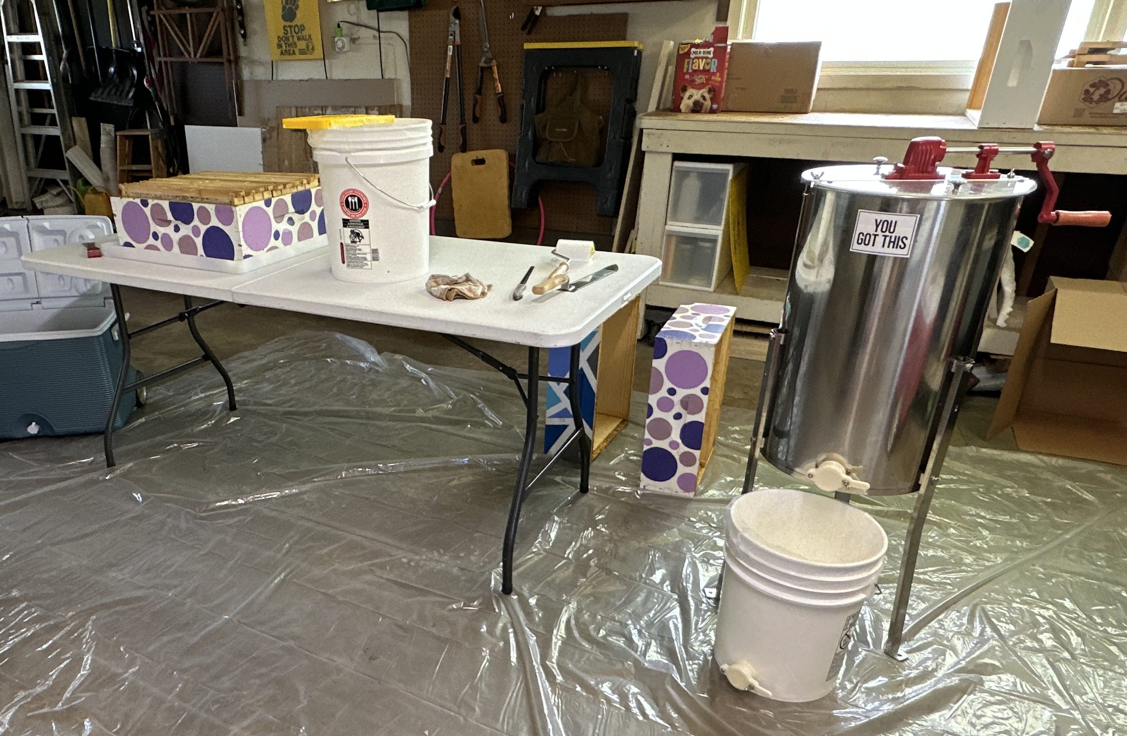 A room with a table, honey extractor, and scattered supplies including a bucket, knife, towel, and disassembled purple beehive box.