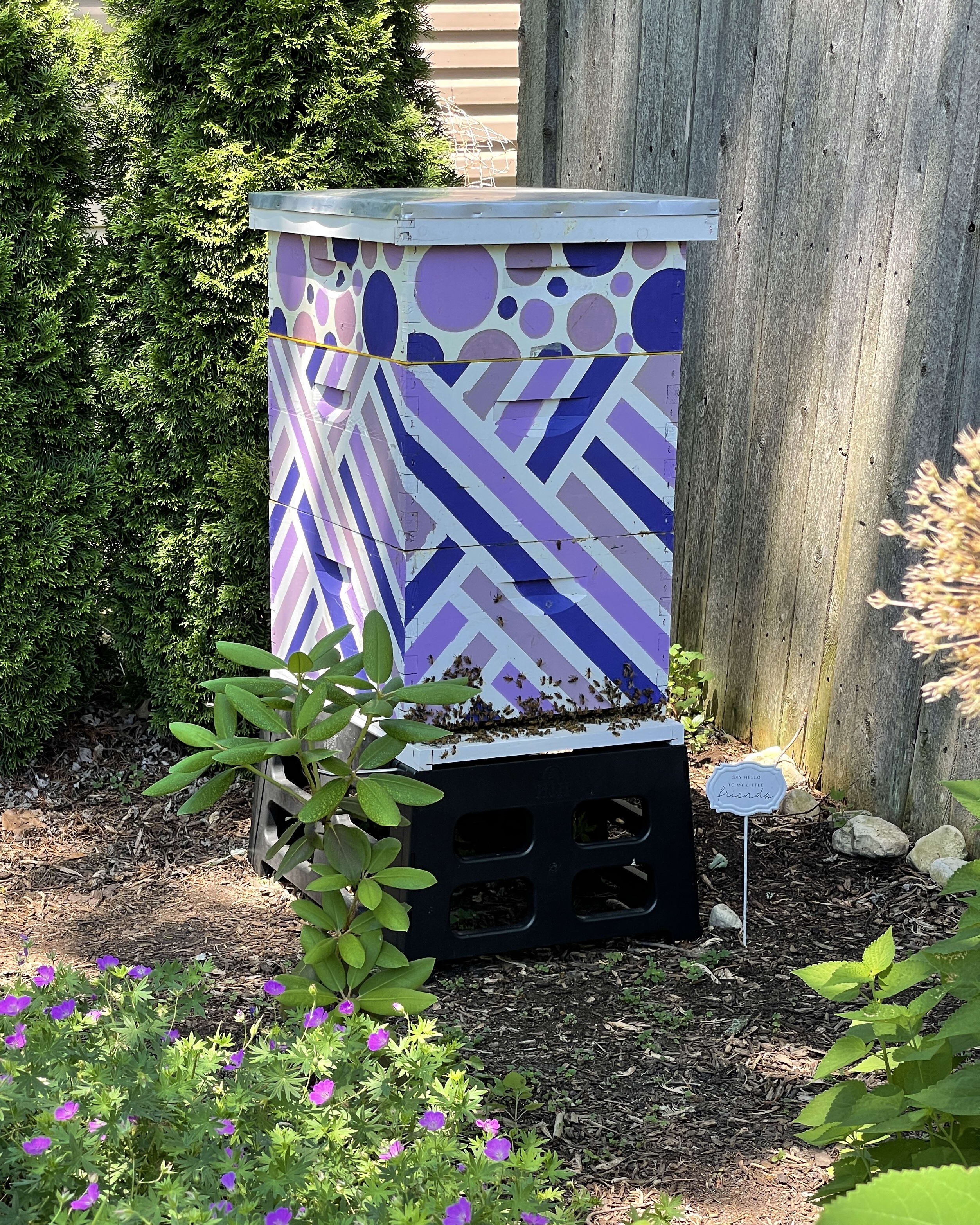 Beehive with a vibrant purple and blue geometric design, placed between shrubs and flowers in a garden.