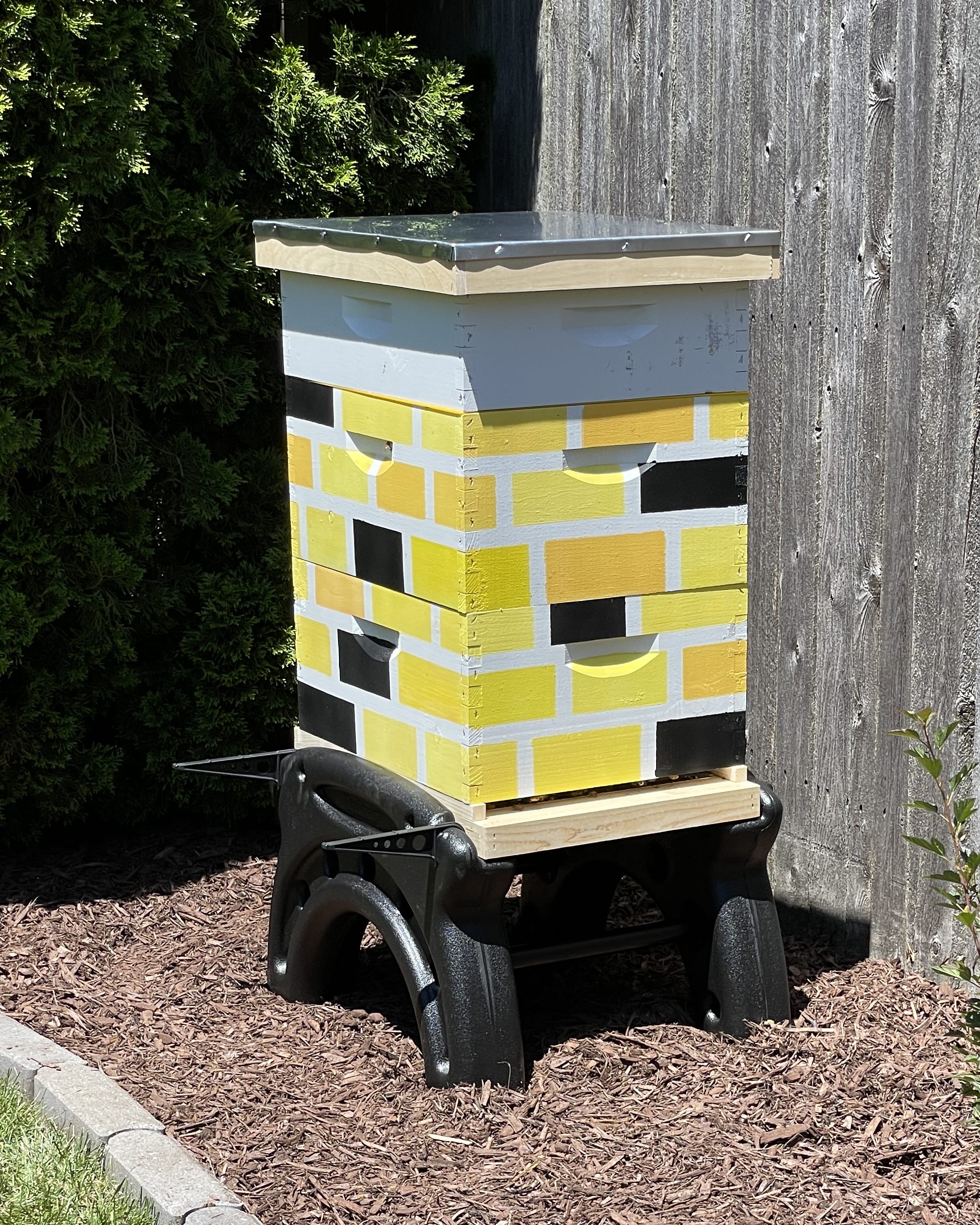 Beehive with a yellow and white pattern, next to a wooden fence and surrounded by mulch.