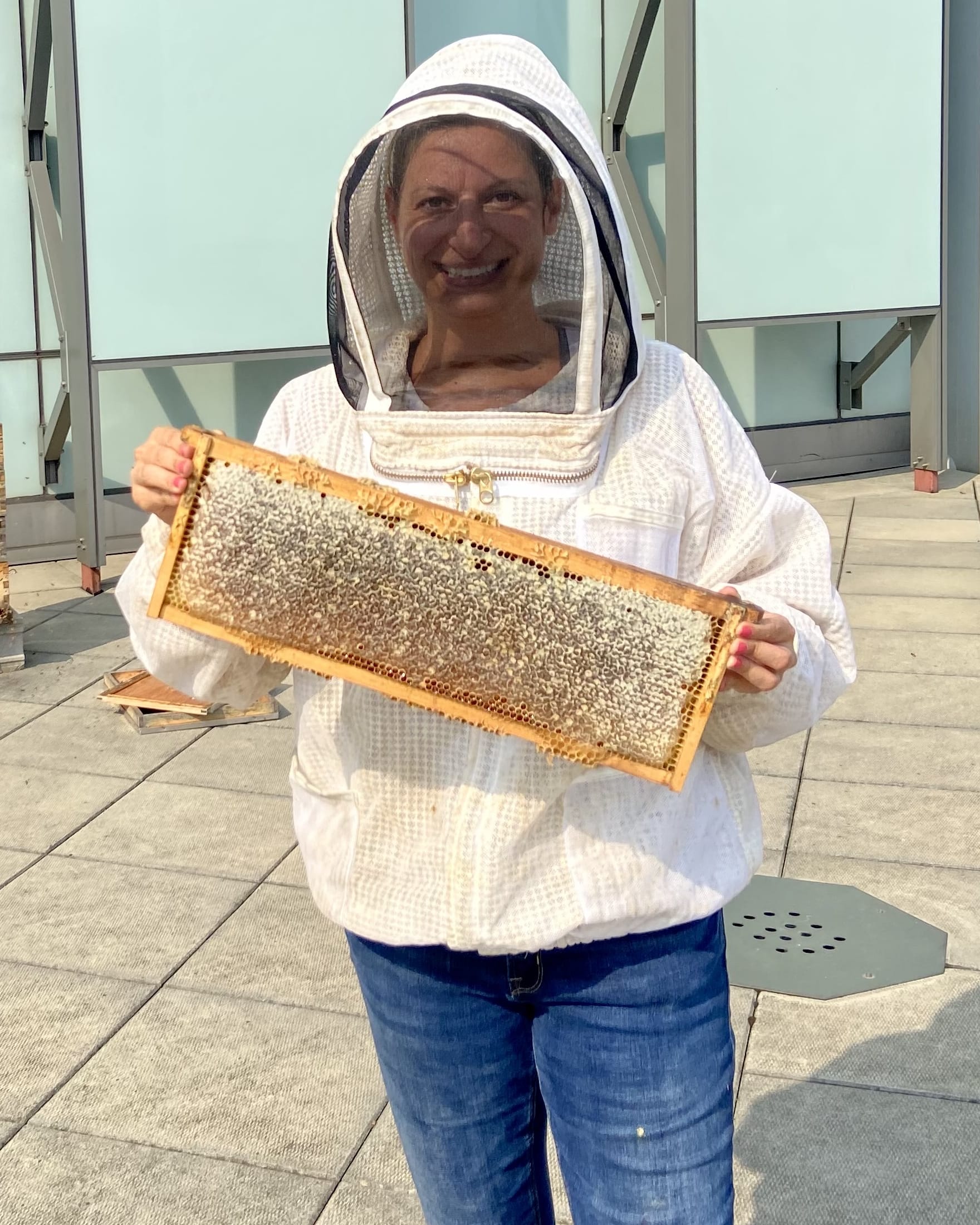 Kate wearing a beekeeping suit and holding up a large frame covered in bees on a sunny day.