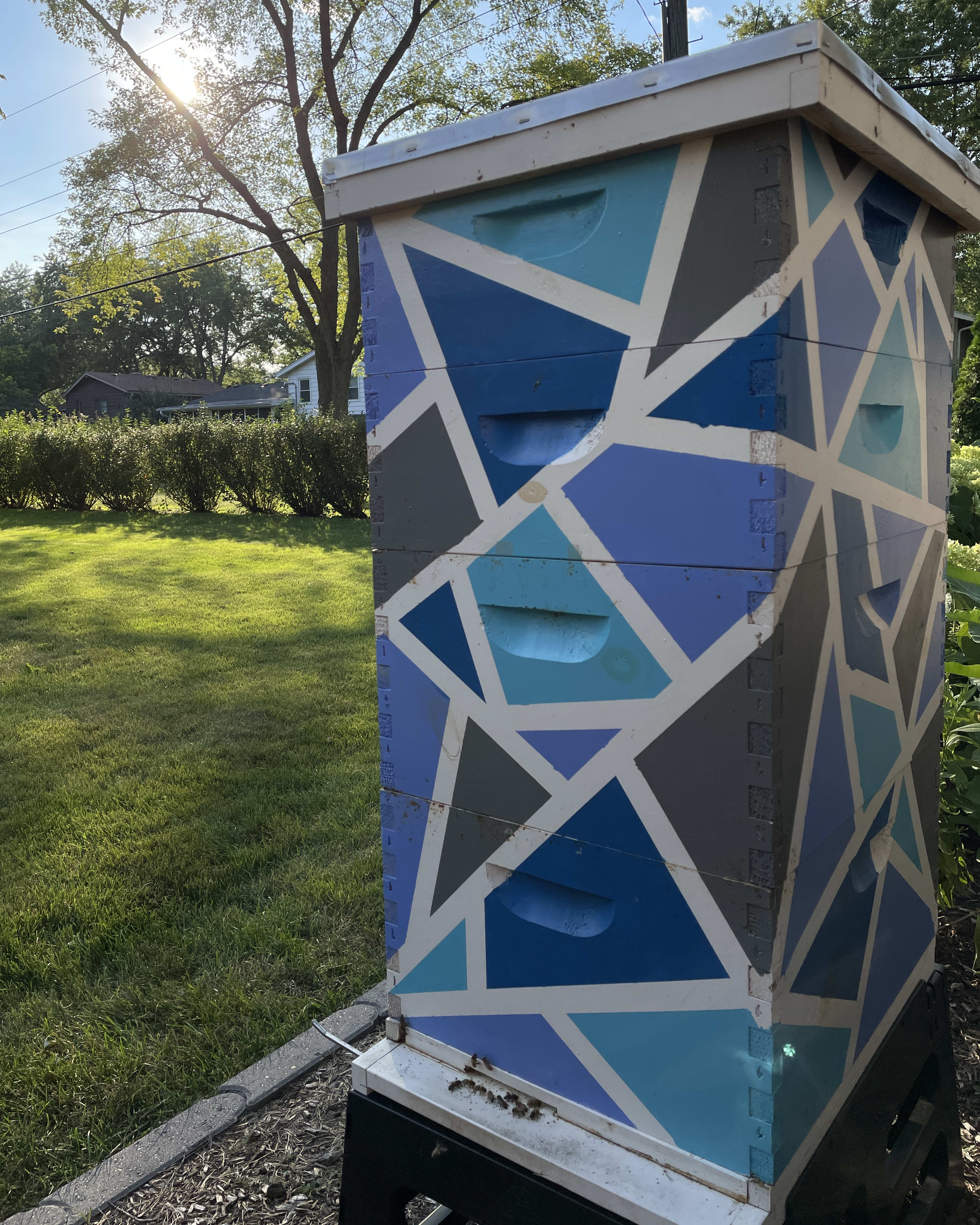 Colorful urban beehive with geometric blue and gray patterns, surrounded by green grass under sunlight.