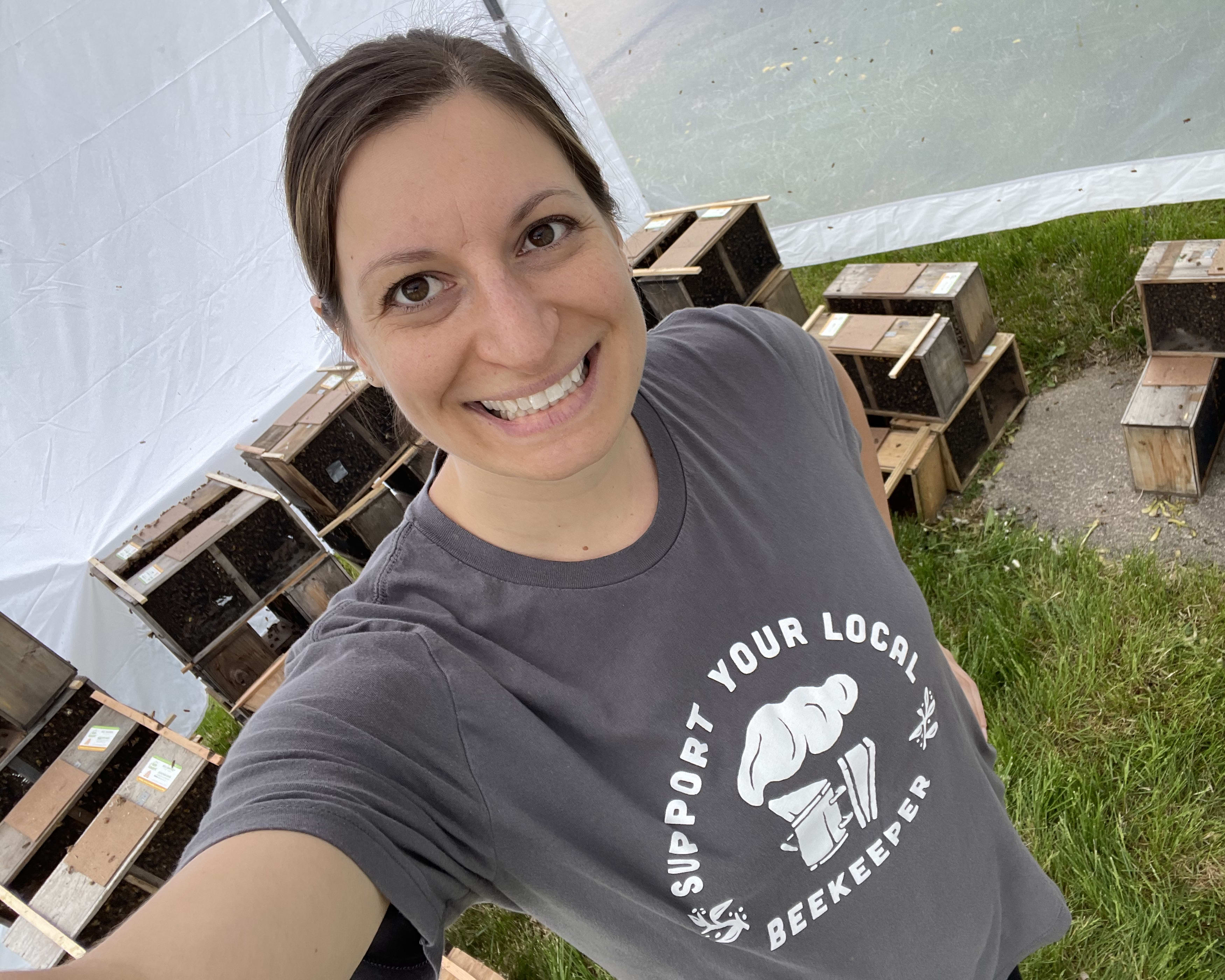 Kate smiling for a selfie, wearing a 'support your local beekeeper' t-shirt, with several bee boxes in the background.