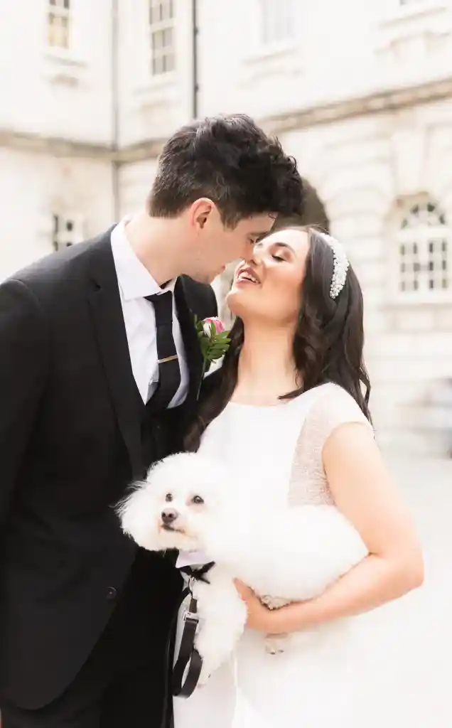 Wedding photo NI of bride kissing groom holding white dog, toy breed