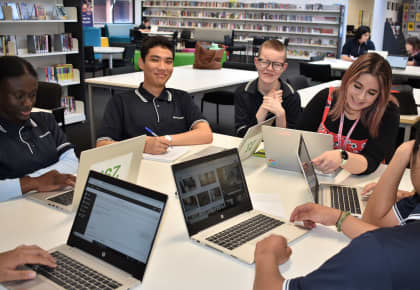 Students sitting at a desk in the library with laptops, pen and paper