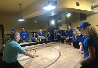 Students from the Anzac tour are standing around a large semi-circle wooden table with numbers etched onto it, a grid and measuring tools used to plot gunfights.