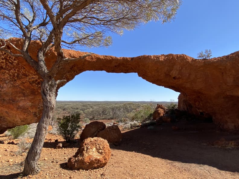 Natural landscape formation in orange rock called 'London Bridge'. A tree stands in front of the bridge on the left hand side and there are some large orange boulders on the ground. Through the arch of the bridge, the view to the horizon is fairly flat with small trees and shrubs. The sky is a brilliant blue.