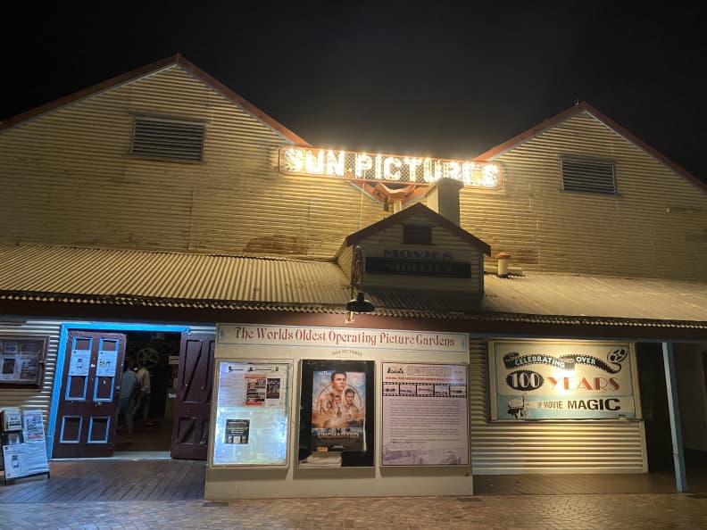 Image shows the outside of the Sun Pictures outdoor cinema gardens in Broome, at night. The building is made from corrugated tin and has a double triangular peaked roof. Between the peaks is an illuminated sign that says "Sun Pictures". The lower half of the building has signs, movie posters and a double wooden door on the left to enter.