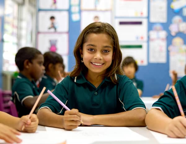 Students at a desk in a classroom, one girl is looking at the camera and smiling