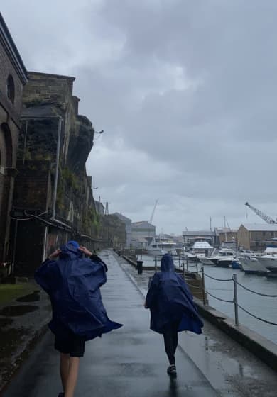 Two students are walking next to the harbour in blue rain poncho's, the sky is full of grey rainclouds