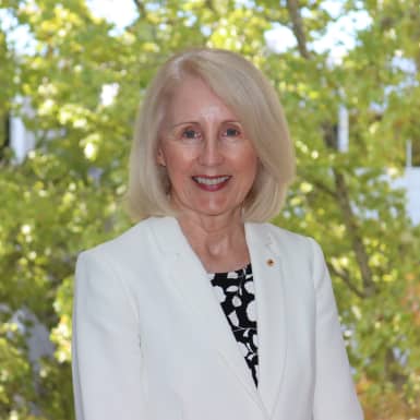 Professor Sandra Harding wearing a white blazer and a black and white patterned blouse with some trees in the background.