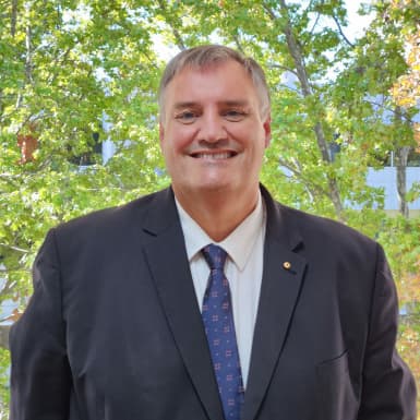 Professor John Williams wearing a black blazer and white shirt with blue tie in front of some trees.