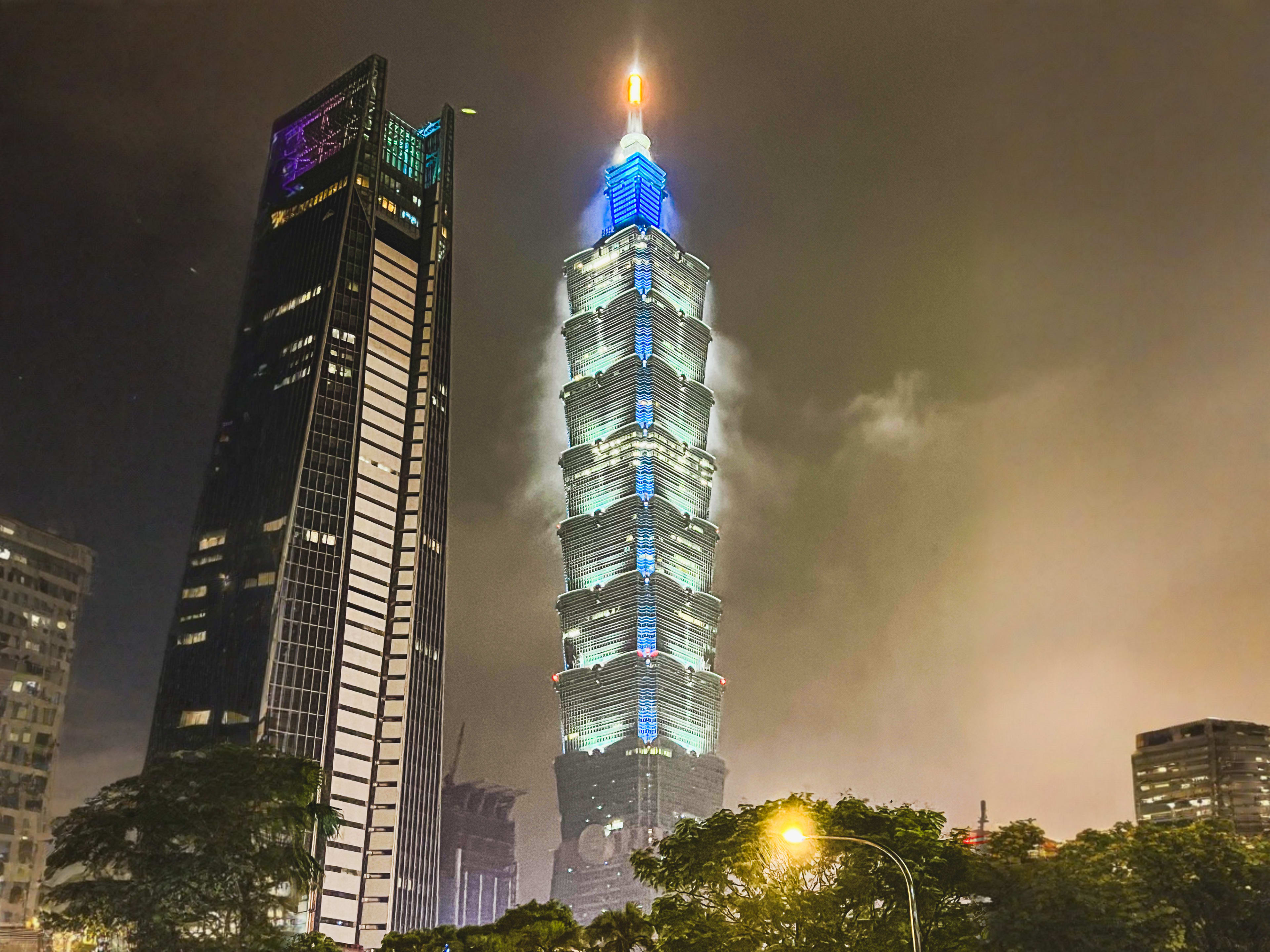 Two tall skyscrapers lit up at night, with the central building brightly illuminated in blue and white lights, rise above trees and clouds—a stunning scene often admired along popular Taiwan cycling routes.
