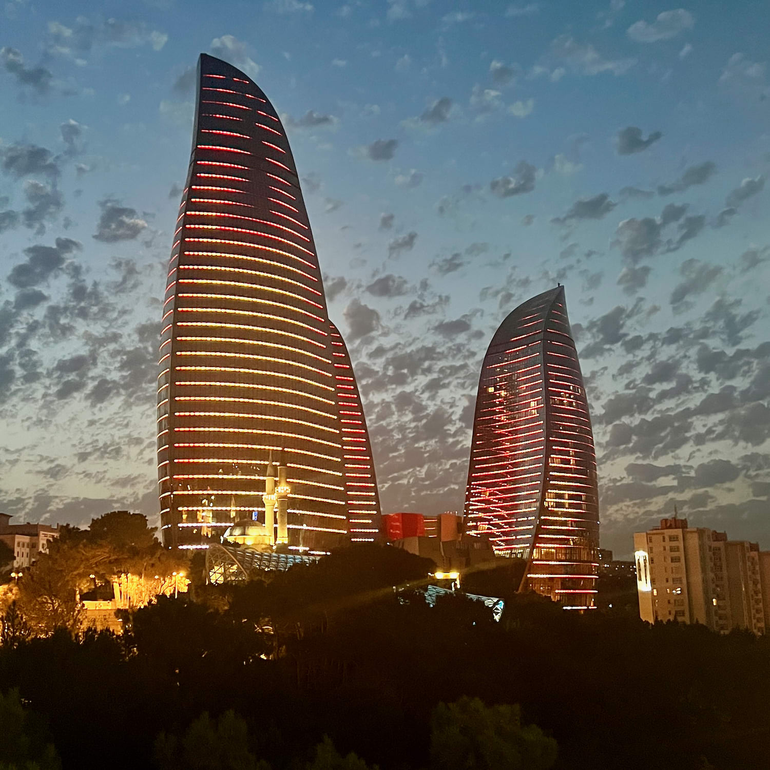 A cityscape of Baku at dusk features two large, uniquely shaped skyscrapers illuminated with red patterns. The sky over the Azerbaijani capital is filled with scattered clouds and a gradient from light blue to deep blue as night approaches.