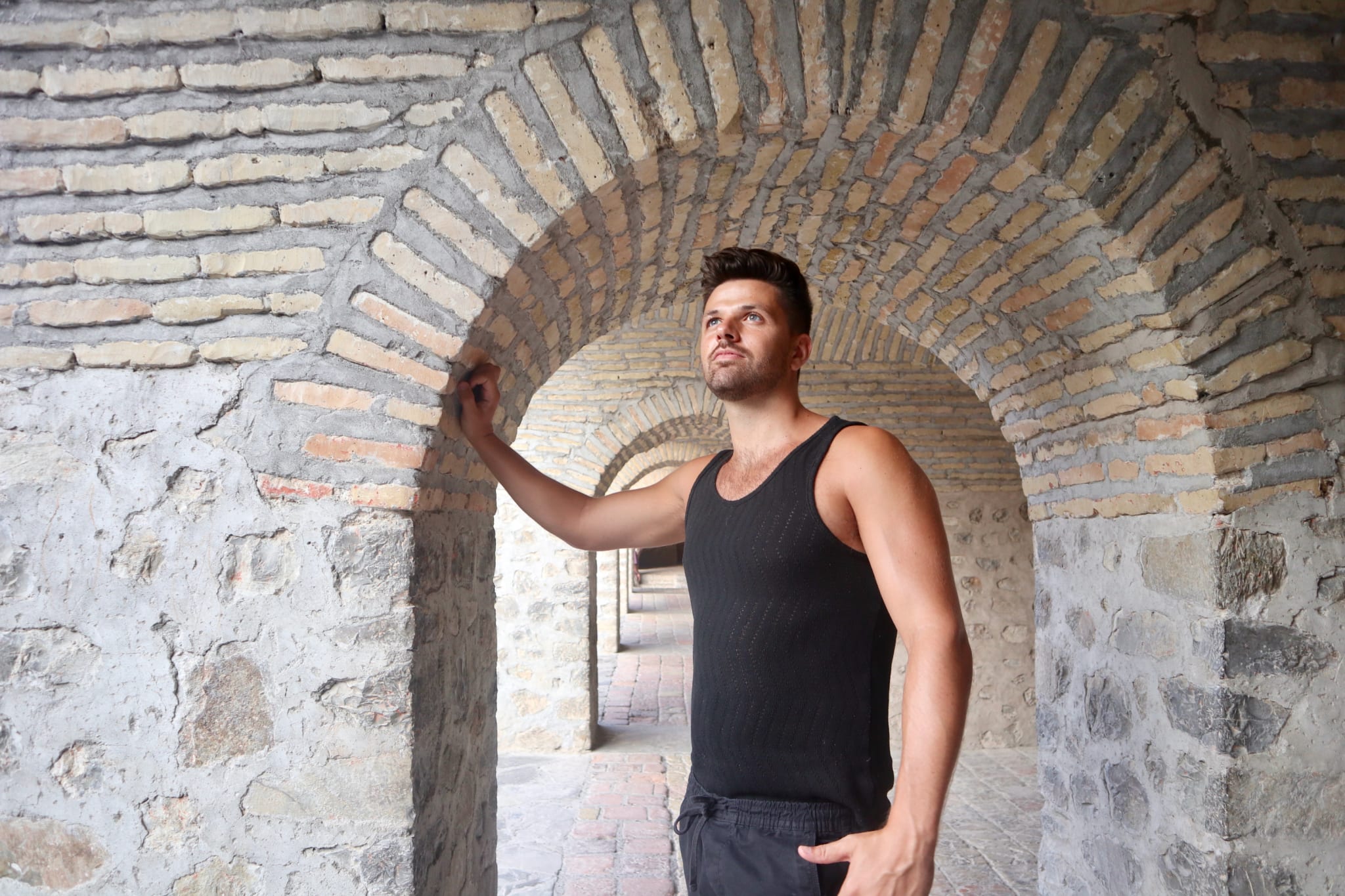 A man in a black sleeveless shirt stands under a stone archway, touching the arch in awe. This historical site resembles Sheki's must-see spots, with its series of arches creating a tunnel effect. Light-colored bricks and stones form the walls, while brick tiles pave the ground—truly one of the many things to do in Sheki for those who appreciate architectural beauty and rich history.