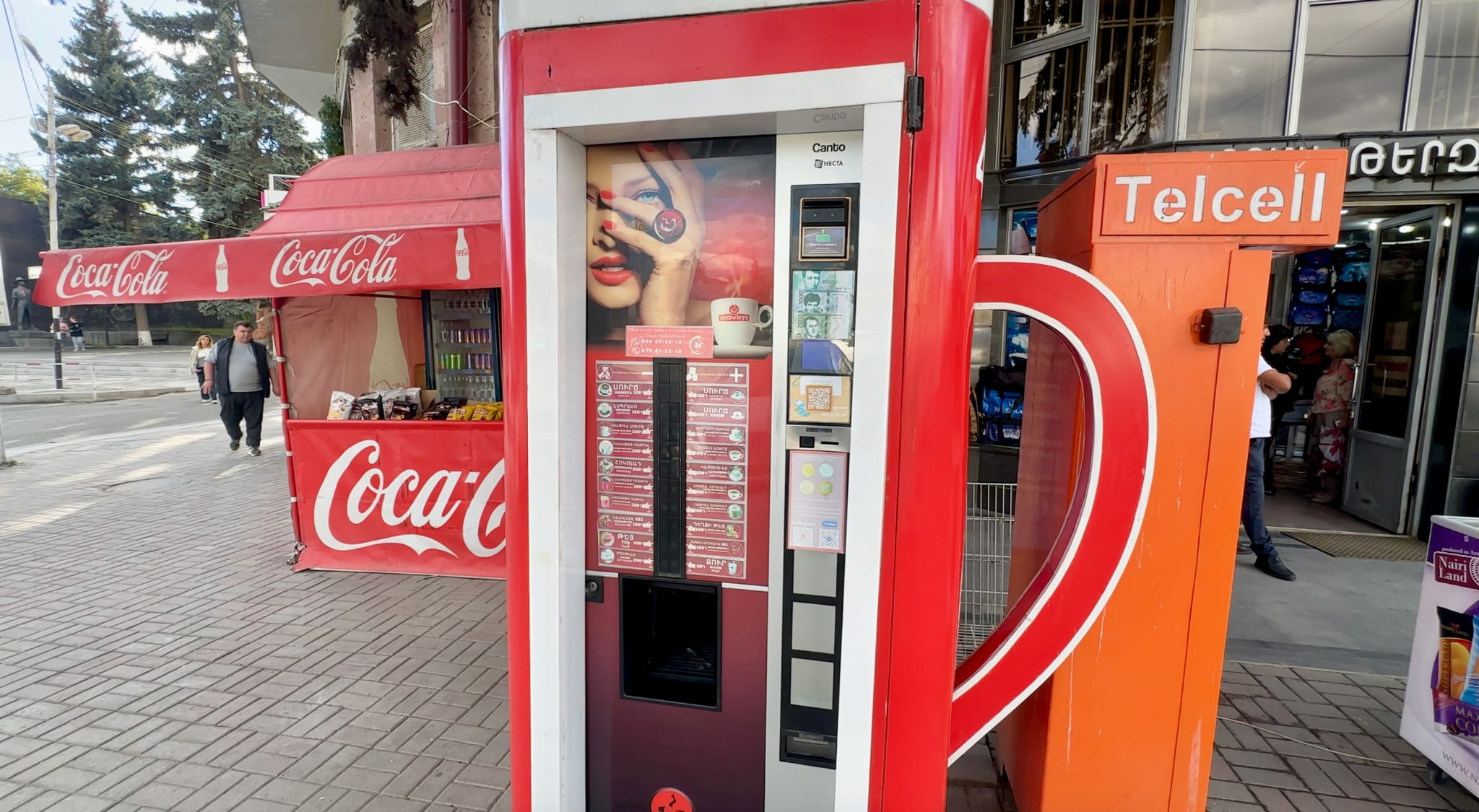 A vibrant red vending machine stands beside a Coca-Cola stand and an orange Telcell box on a bustling Gyumri sidewalk in Armenia. It's one of the many intriguing sights and gyumri things to do while exploring the city's unique blend of modernity and tradition.