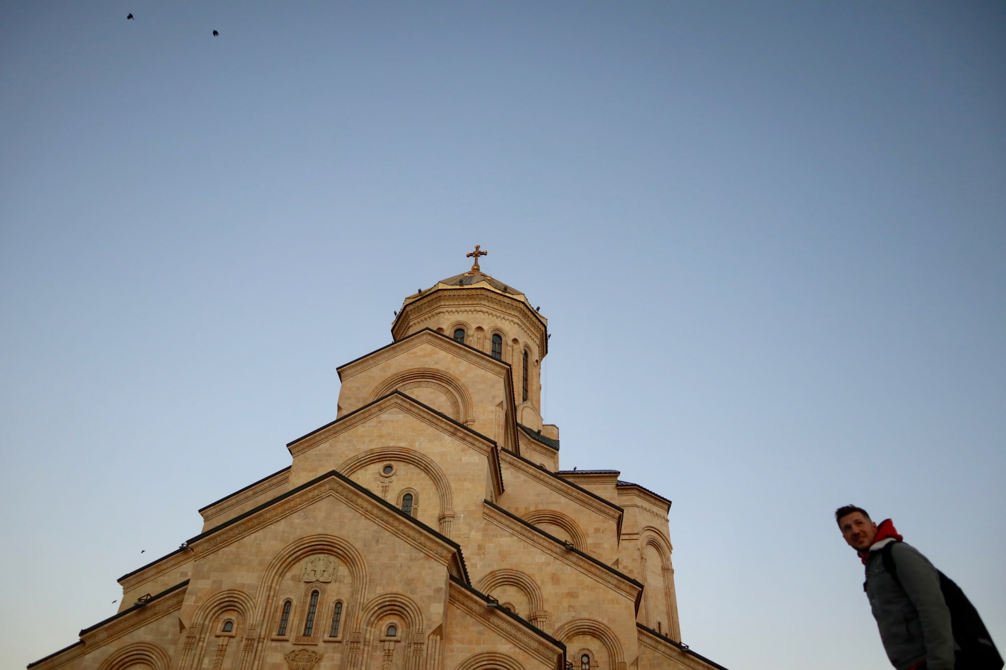 A man strolls near a large beige cathedral with a prominent dome and cross, basking in the clear sky. This stunning architectural gem is among the must-see spots for anyone exploring Georgia's travel highlights. If you're wondering, 'is Georgia worth visiting?' this breathtaking scene provides an emphatic yes, showcasing the country's rich cultural heritage and beauty.