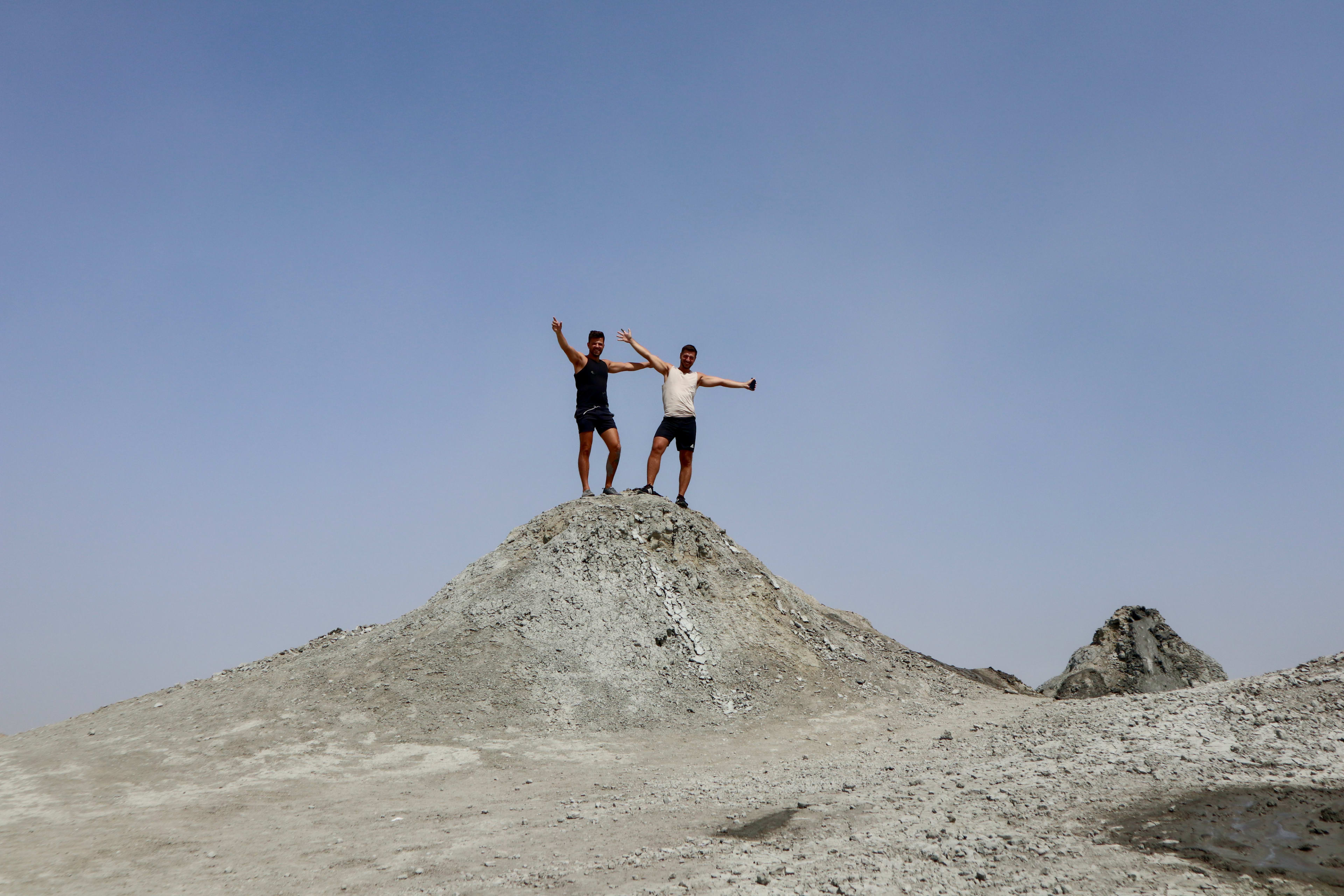 Two people standing on top of a rocky mound with arms raised in celebration. The sky is clear and blue, creating a stark contrast with the grey, barren landscape. Both are dressed in casual, summer clothing. Plan your own trip to Azerbaijan for an adventure like this!