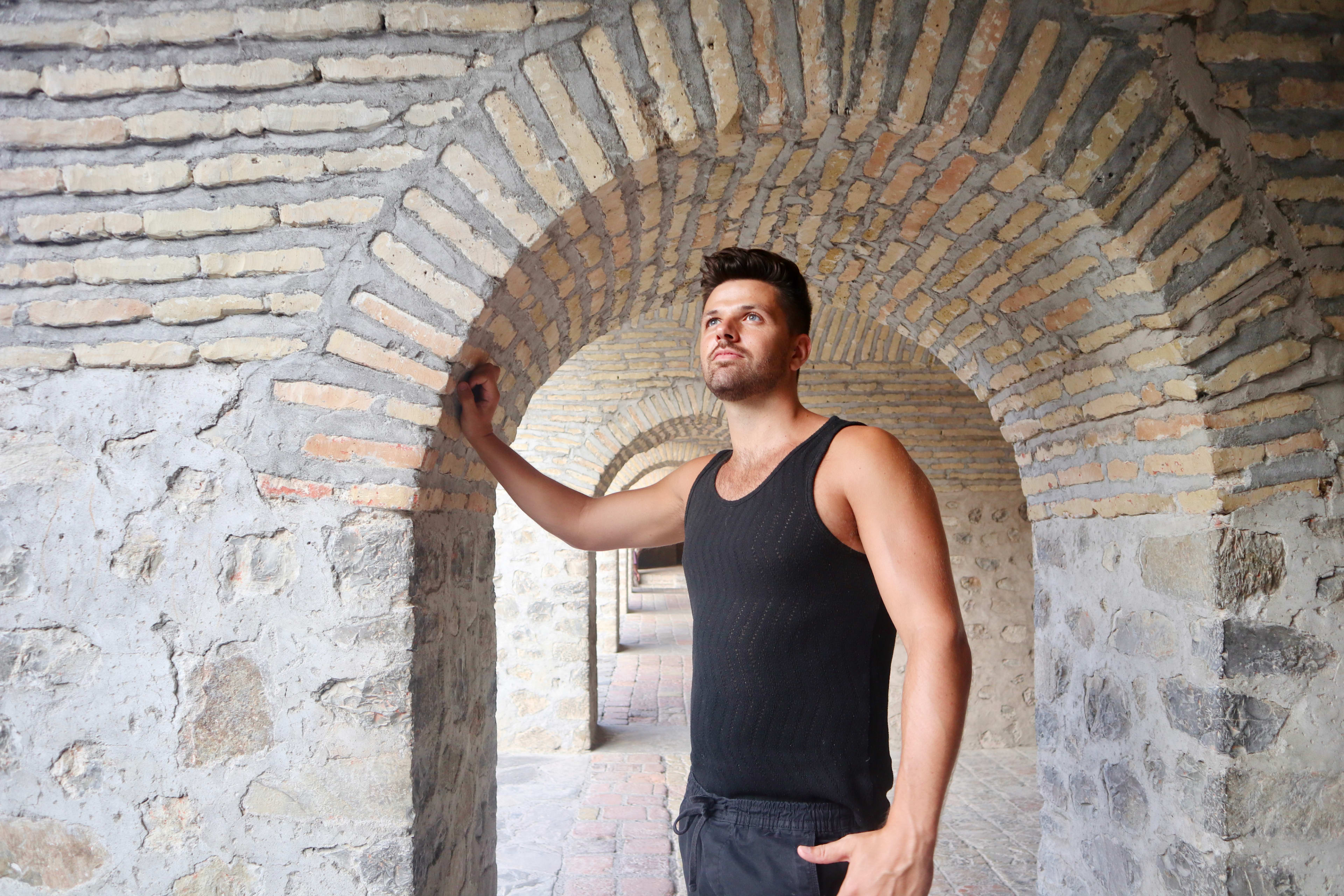 A man in a black tank top stands under an arched stone hallway, touching the archway with his left hand. He looks slightly to the side with a thoughtful expression. The setting has a historical and architectural ambiance reminiscent of the Silk Road era, with brick and stone walls.