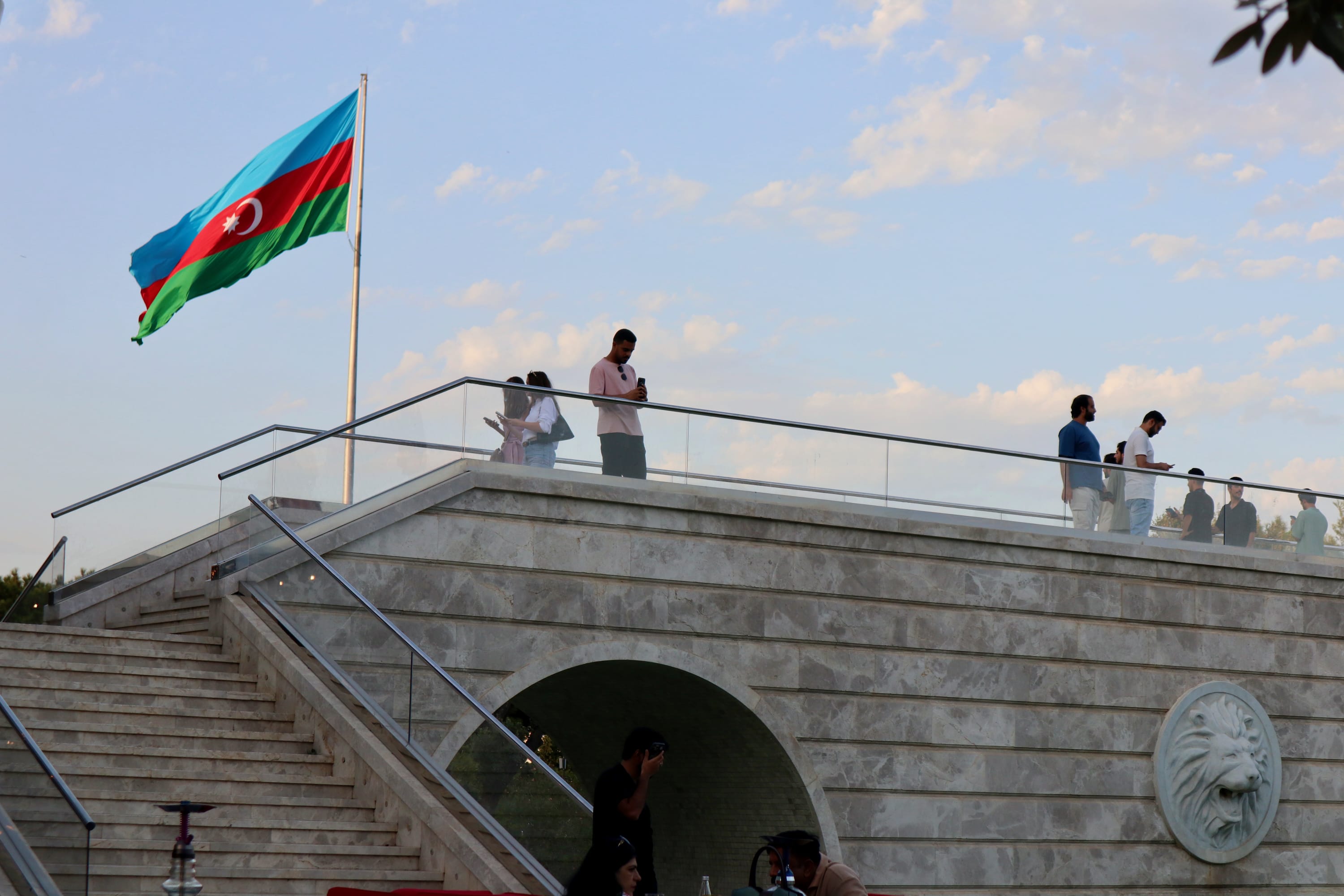 People visiting Baku stand on a marble bridge with a large Azerbaijani flag waving in the breeze. The bridge features a lion face design on the side. A clear blue sky with scattered clouds serves as the backdrop, highlighting Azerbaijan's vibrant charm.