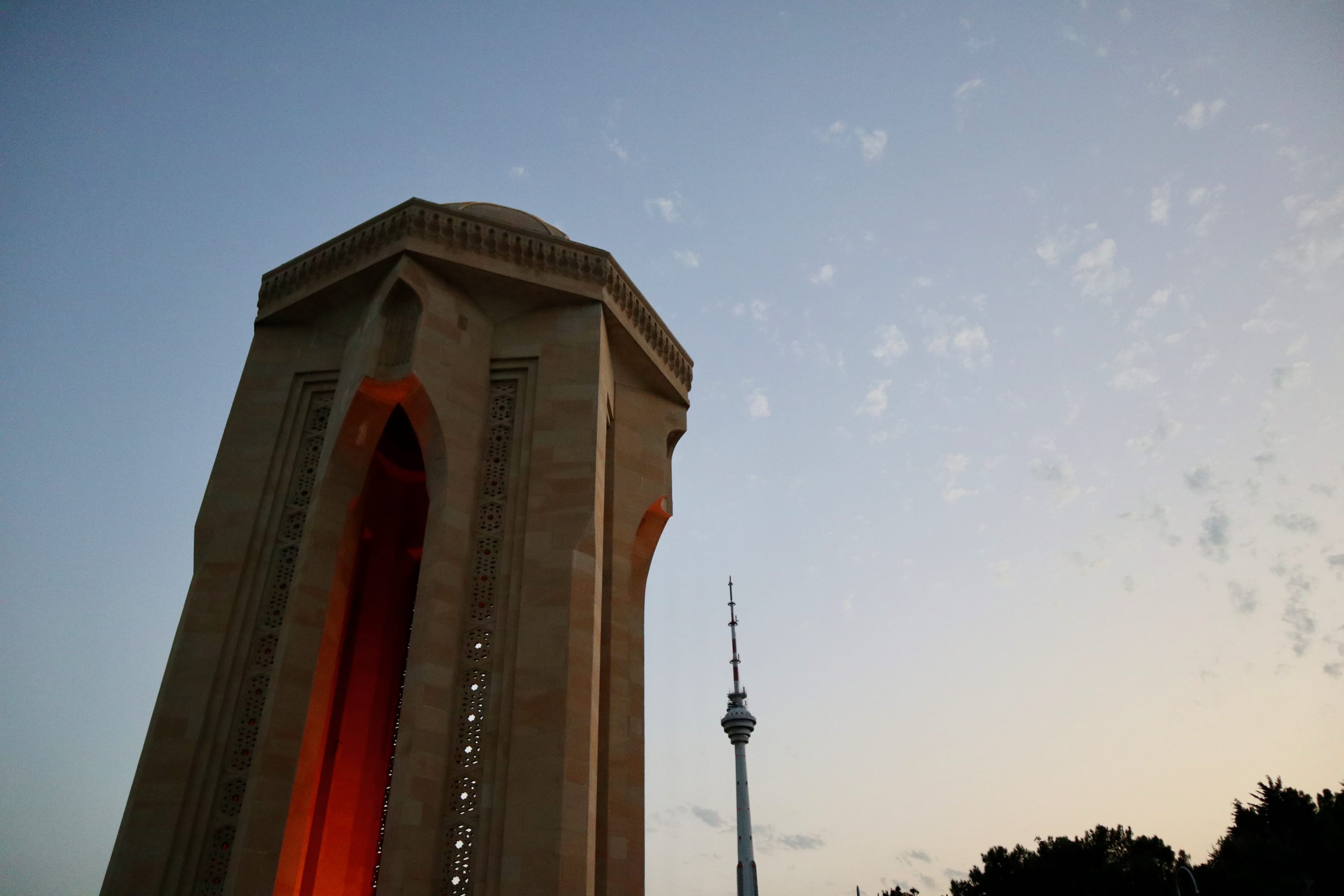 A tall, ornate tower with intricate carvings is prominently featured on the left, set against a dusky sky over Baku, the Azerbaijani capital. Another slender tower with an antenna rises in the background on the right. Trees gracefully frame this enchanting scene below.