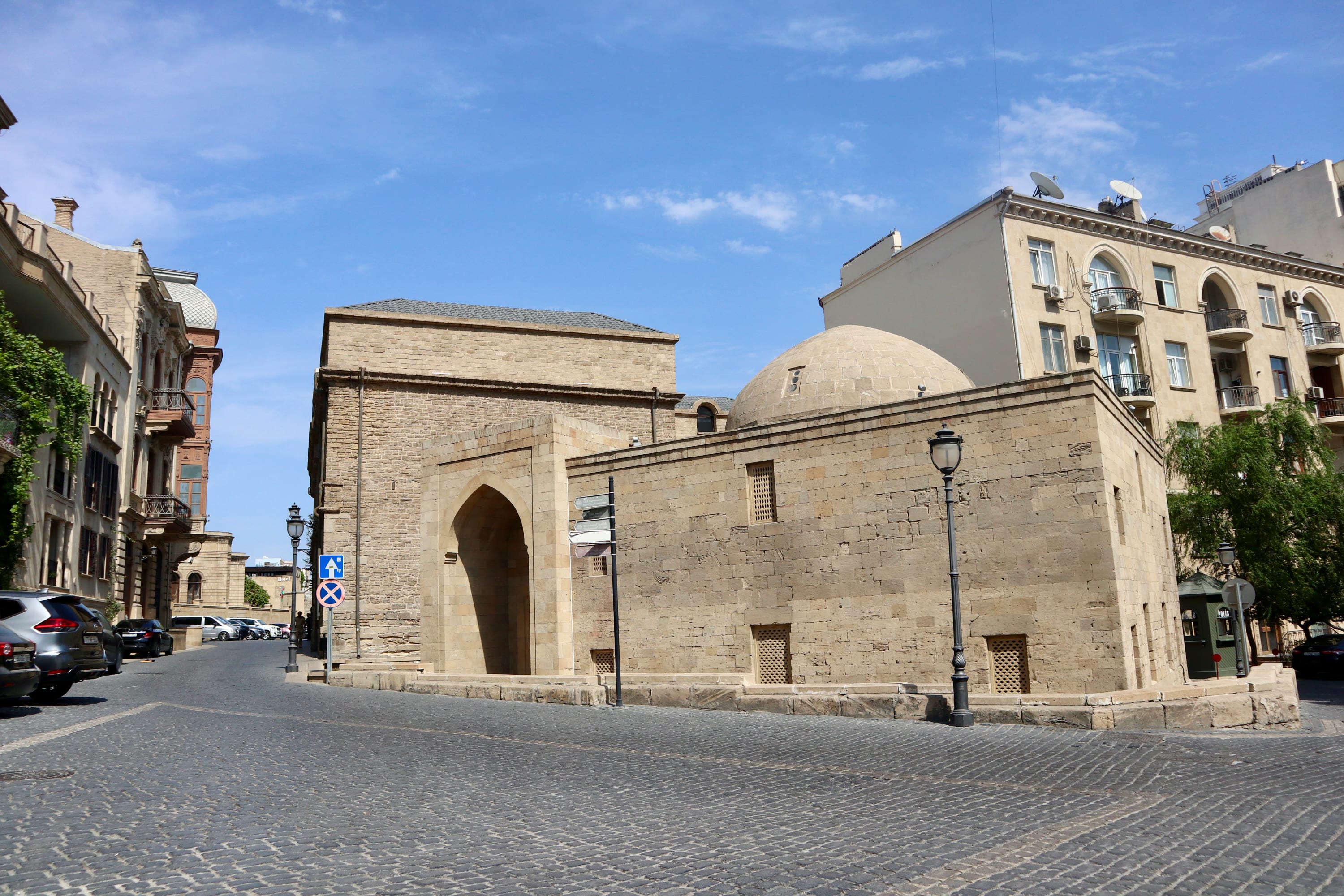Nestled in the heart of Baku, Azerbaijan, this historical stone building with an arched entrance and dome graces a cobblestone street. Trip planning is a joy with its charming surroundings of street lamps and cars, all under a clear blue sky framed by additional buildings.