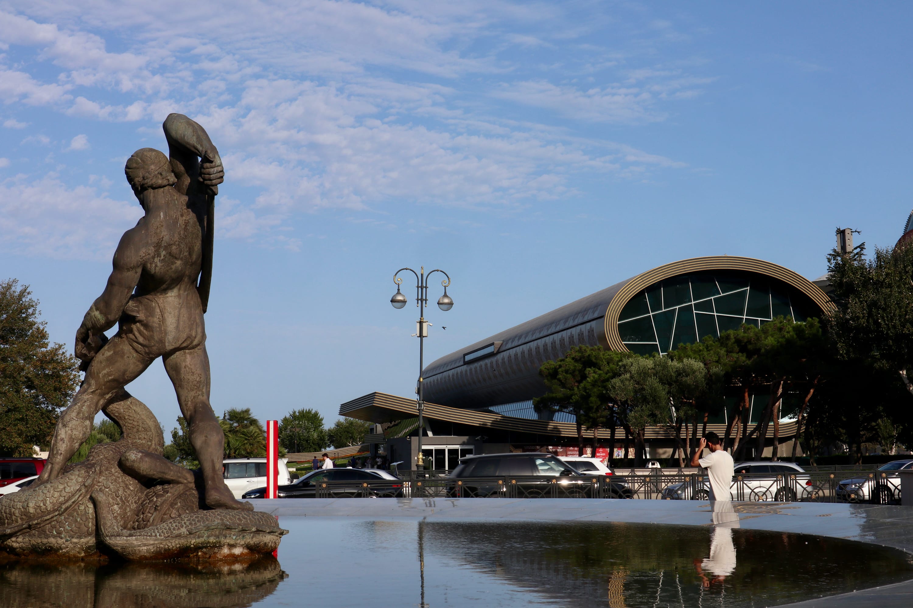 A statue of a man and sea creature stands majestically in front of a modern cylindrical building, capturing the essence of Baku. Nearby, a visitor takes a photo beside the fountain, with parked cars and trees silhouetted against Azerbaijan's clear blue sky.