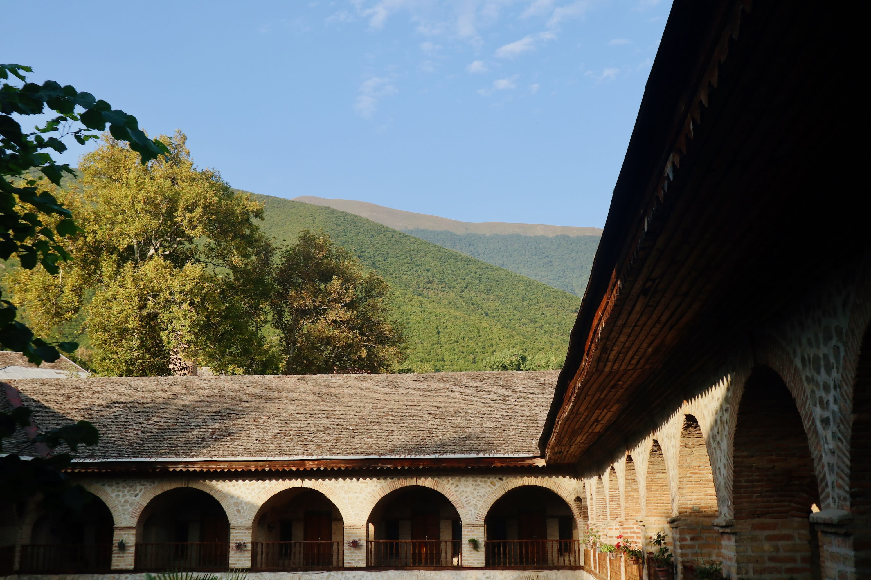 An outdoor courtyard with a series of arches supports a wooden roof. Surrounding the courtyard is a lush landscape of trees and a mountain in the background under a blue sky, reminiscent of Sheki. The architecture appears to be historical, featuring stone and brick elements.