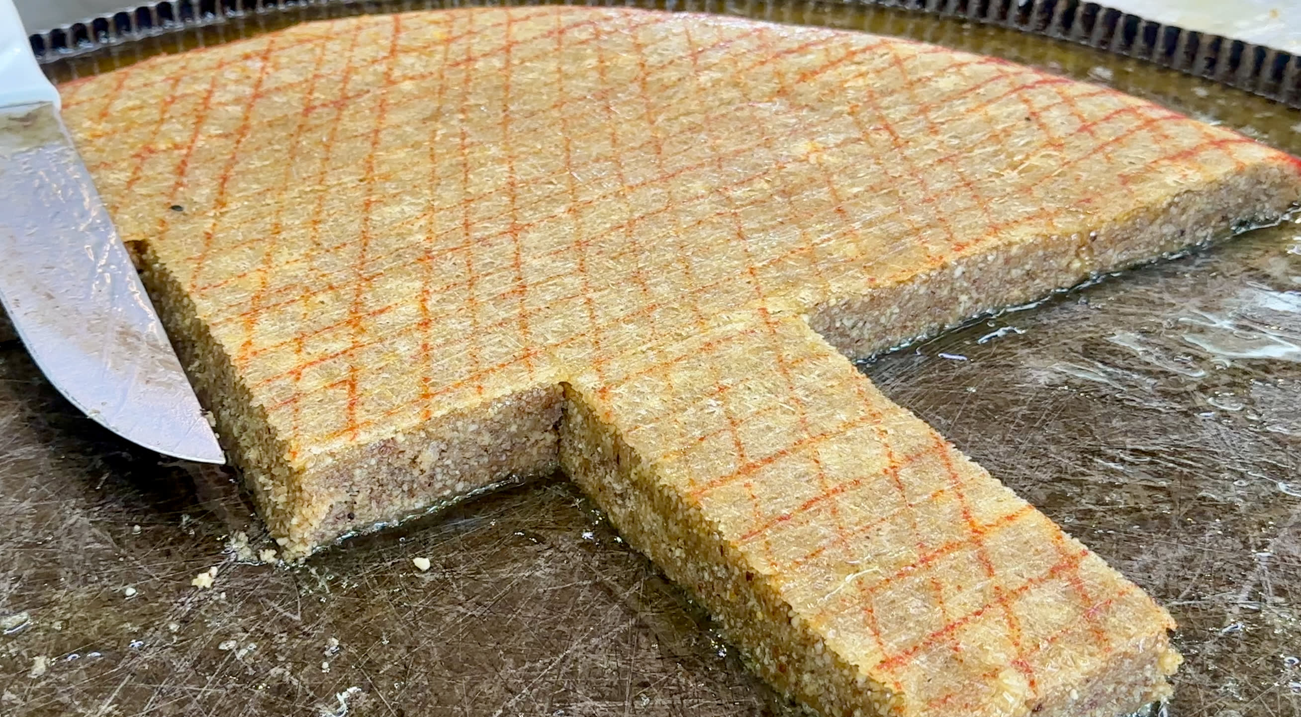 A large circular tray of sheki halva is shown with a triangular portion cut out. The golden-brown dish, resembling the historical patterns found in Sheki's must-see spots, displays a grid pattern on top from knife scores. A knife rests beside the missing piece, highlighting its textured surface. When visiting Sheki, one of the many things to do is indulging in traditional Azerbaijani cuisine like this baklava or halva and exploring its rich culinary heritage.