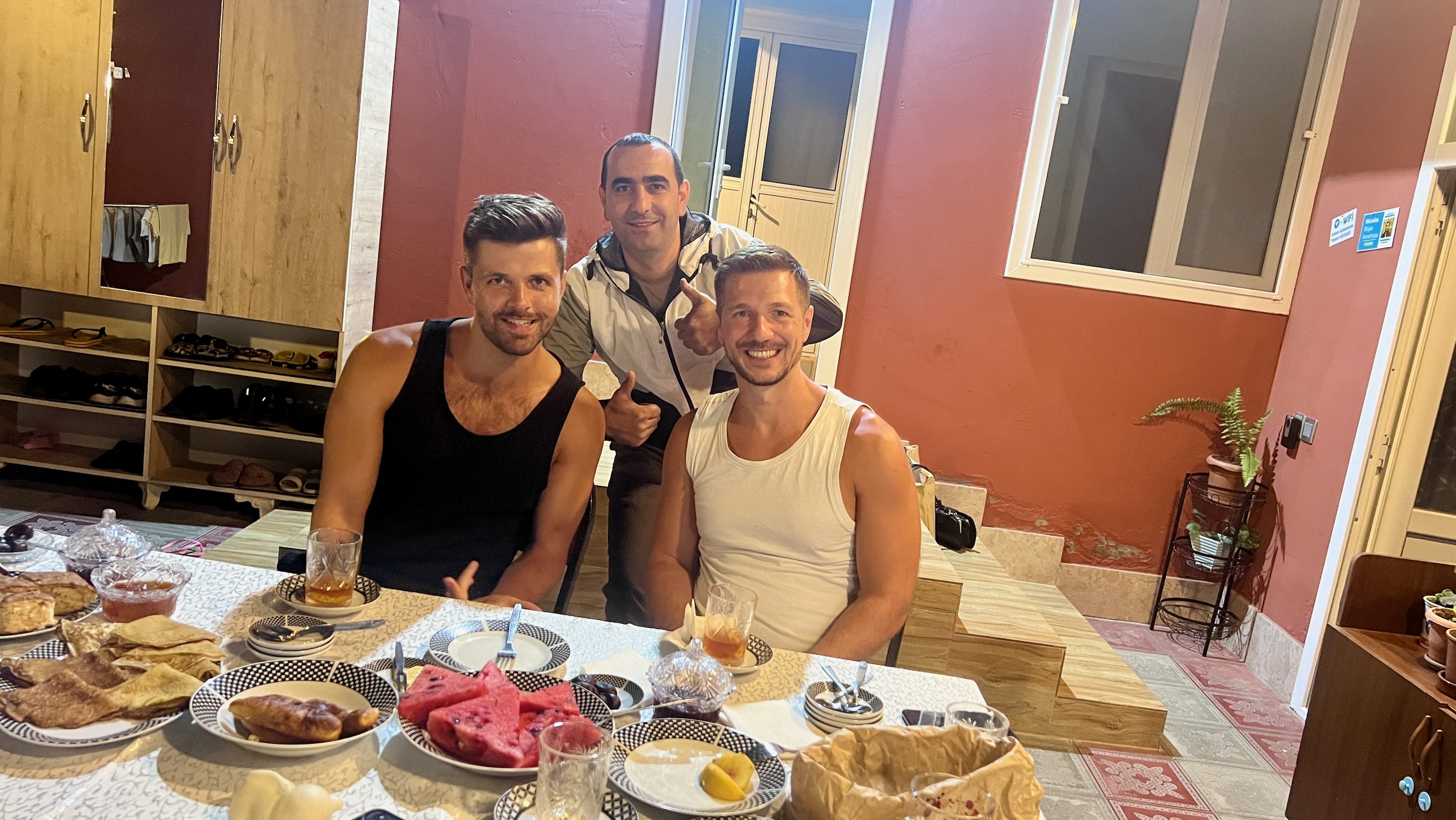 Three men are seated at a dining table laden with food, including watermelon and bread. They are smiling, with two sitting and one standing behind them. The room's red walls evoke the rich history of Sheki, while shelves with shoes in the background add to the casual and warm atmosphere.