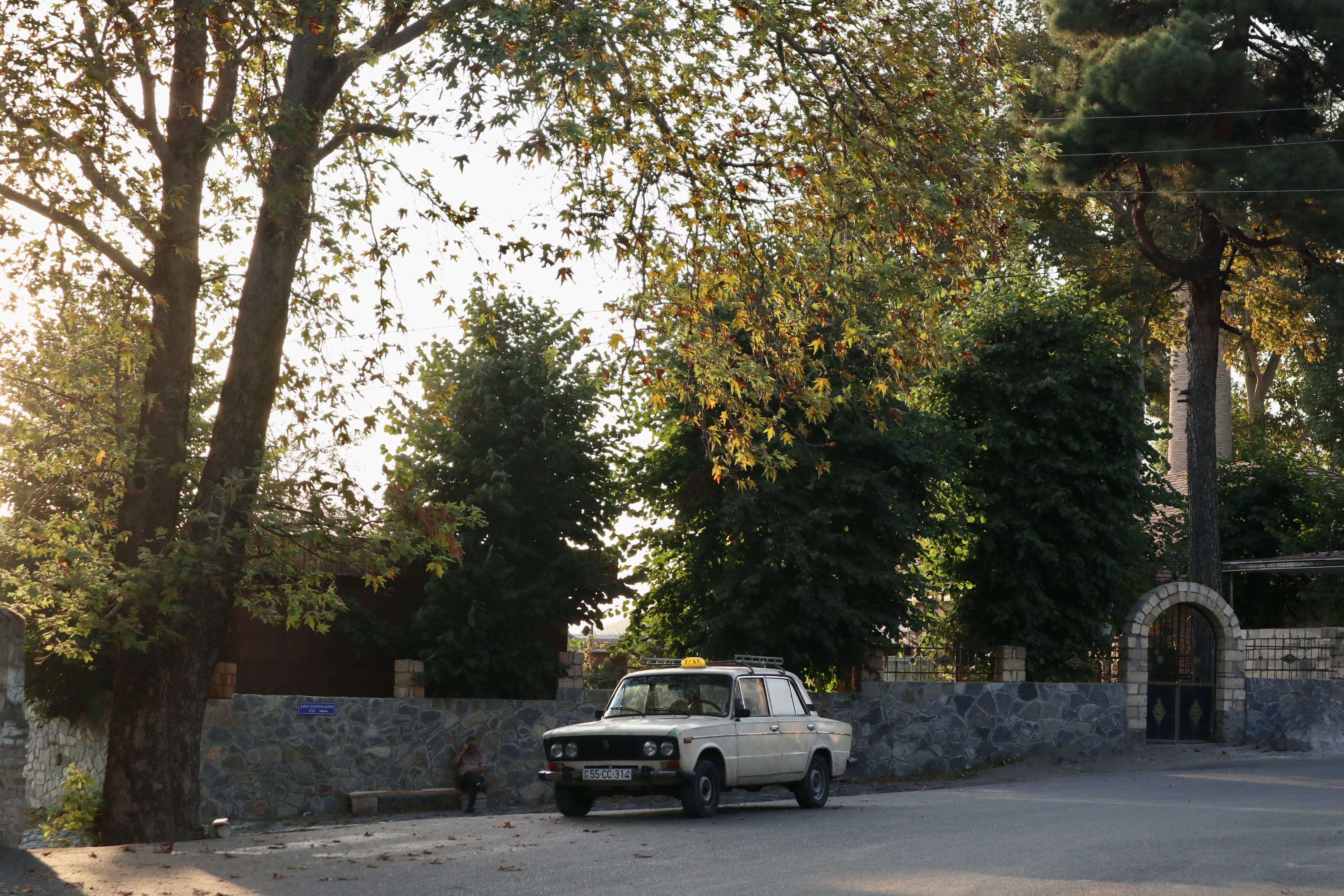 A vintage cream-colored car is parked on a quiet tree-lined street, reminiscent of Sheki's historic charm, where there are many things to do in Sheki. The sun sets, casting a warm glow over the scene. Stone walls and a gate, partially hidden by lush foliage, enhance the peaceful and nostalgic atmosphere.