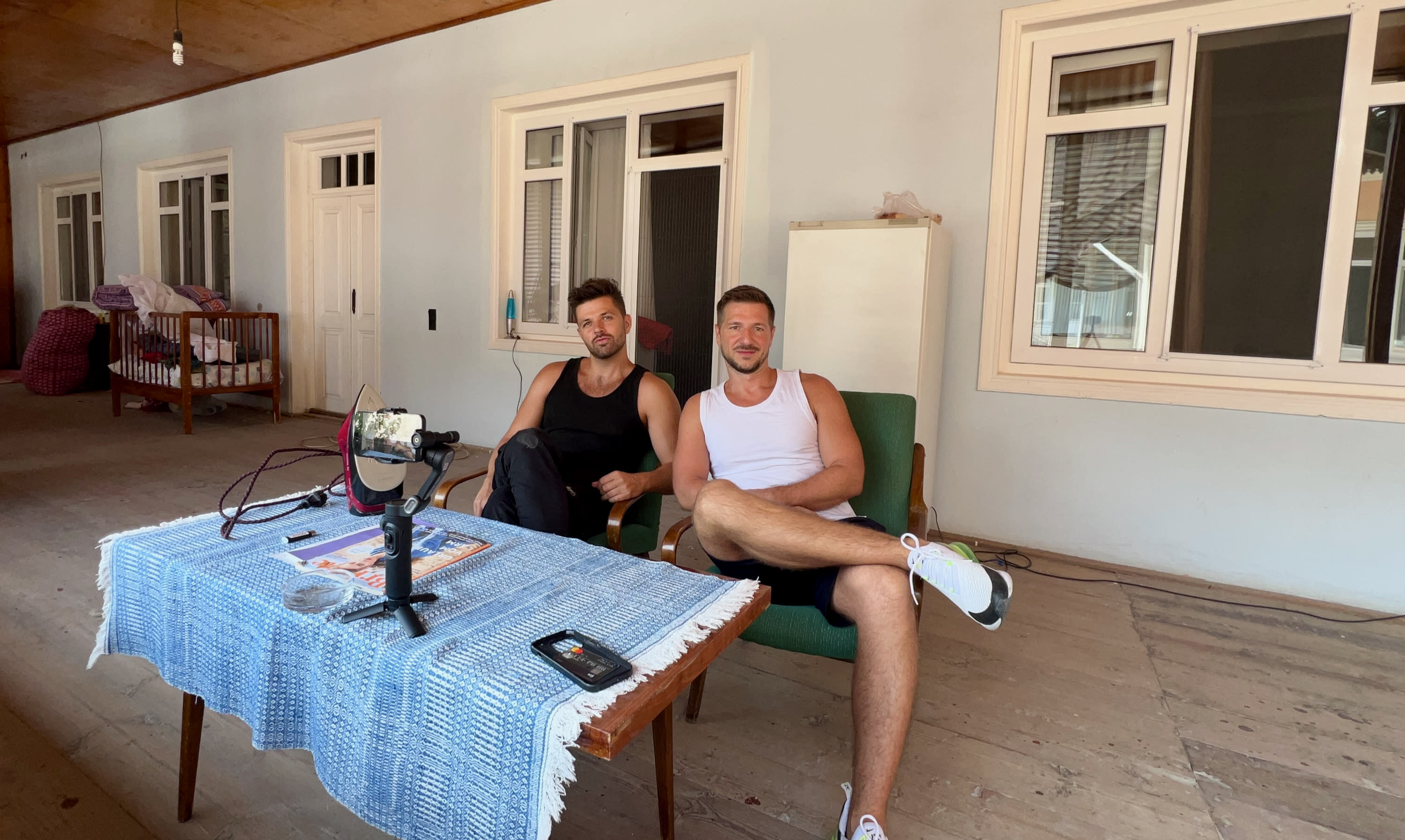 Two men sit on a green chair and armchair on a porch with white-framed windows, reminiscent of Sheki's charming old architecture, creating an inviting atmosphere for visitors exploring Sheki. A table adorned with a blue lace cloth holds electronic equipment and a phone. Both wear tank tops and shorts, appearing relaxed in the outdoor setting, embodying the laid-back vibe often associated with this vibrant destination.