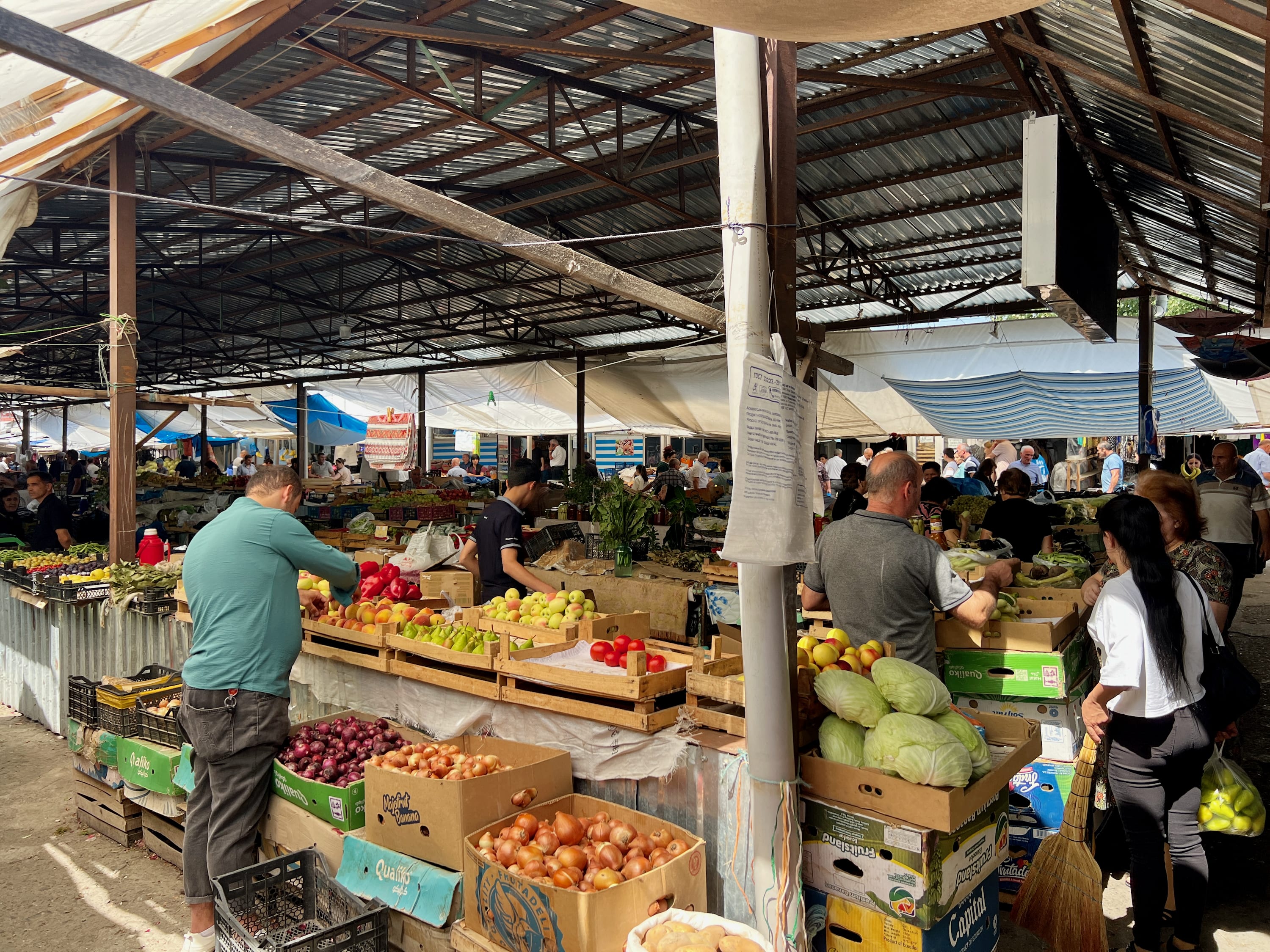 Visitors looking for things to do in Kish, Azerbaijan, can explore its lively outdoor market. Here, people wander through stalls filled with vibrant fruits and vegetables under a metal roof, capturing the essence of local culture. This bustling scene offers a perfect snapshot of Kish's everyday life, making it a must-see on any travel itinerary.