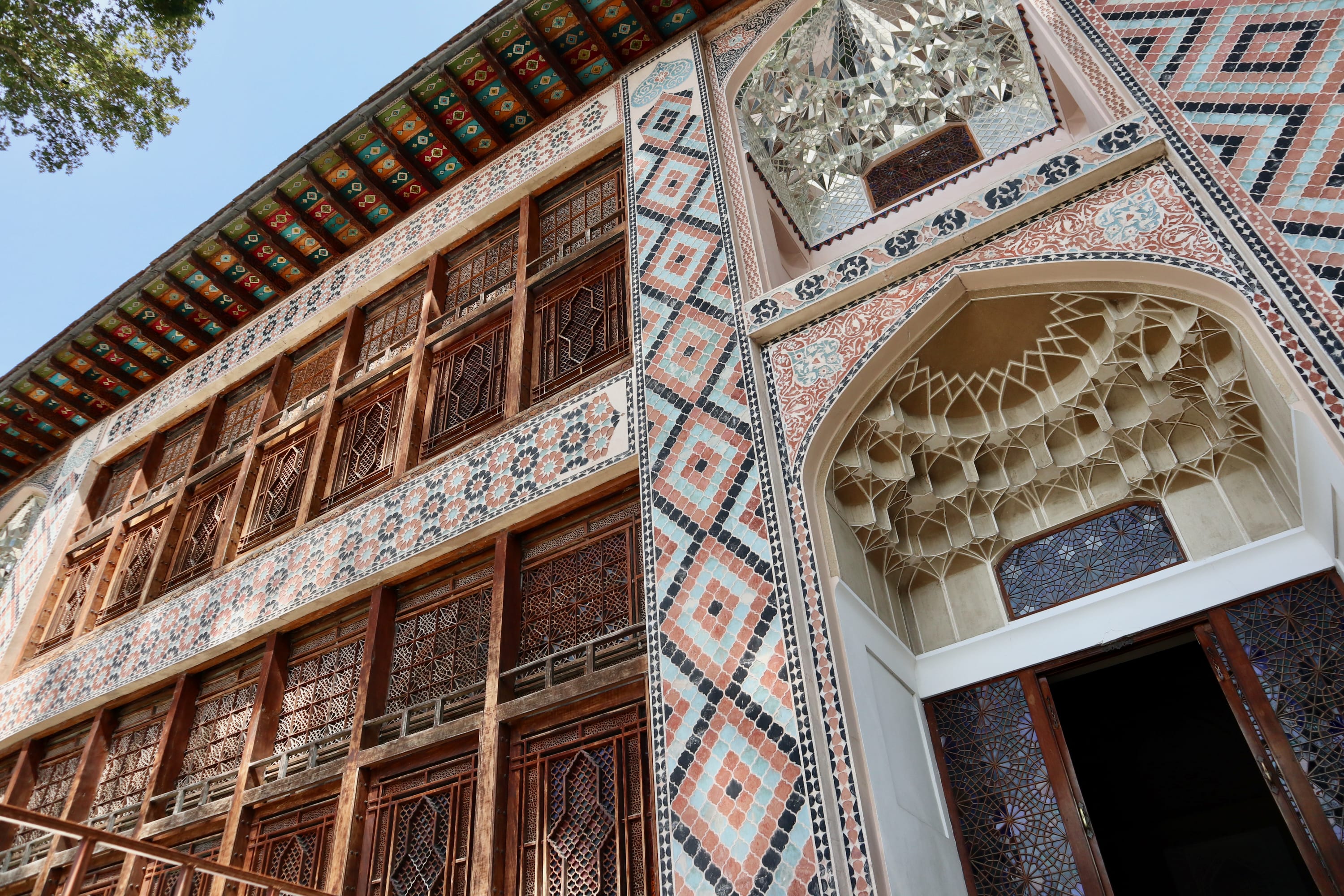The image showcases a historic building in Sheki, adorned with intricate geometric tile patterns in blue, orange, and white. Wooden lattice windows line the facade, while a large arched entrance boasts ornate stonework. A tree stands to the left under a clear sky, inviting viewers on a journey through time. For those seeking things to do in Sheki, exploring such architectural marvels provides an enriching glimpse into the region's rich cultural and historical tapestry.