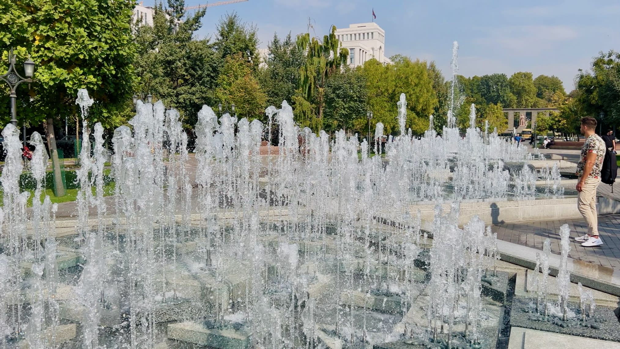 In a vibrant park in Yerevan, a dazzling fountain with multiple water jets dances gracefully amidst lush trees and towering buildings. To the right, someone engages with a gay travel guide, eager to uncover local LGBTQ+ spots and vibrant events like the annual Gay Yerevan festival that celebrates diversity in this welcoming city.