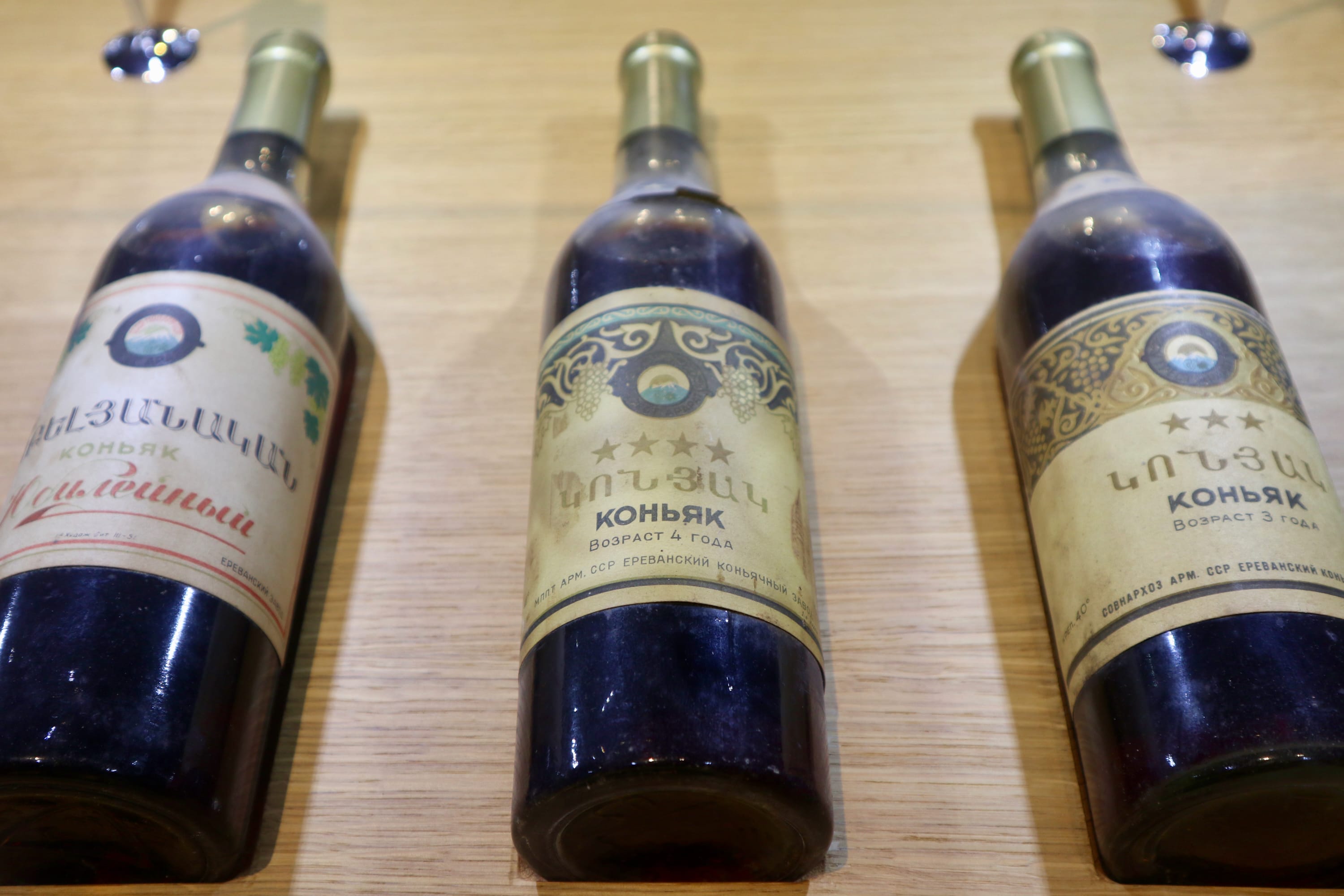 Three bottles of Armenian brandy, each bearing a distinctive Cyrillic label and gold foil cap, are displayed on a wooden surface. These treasures from Yerevan capture the spirit of local culture, with varying label designs that celebrate traditional craftsmanship and rich heritage.