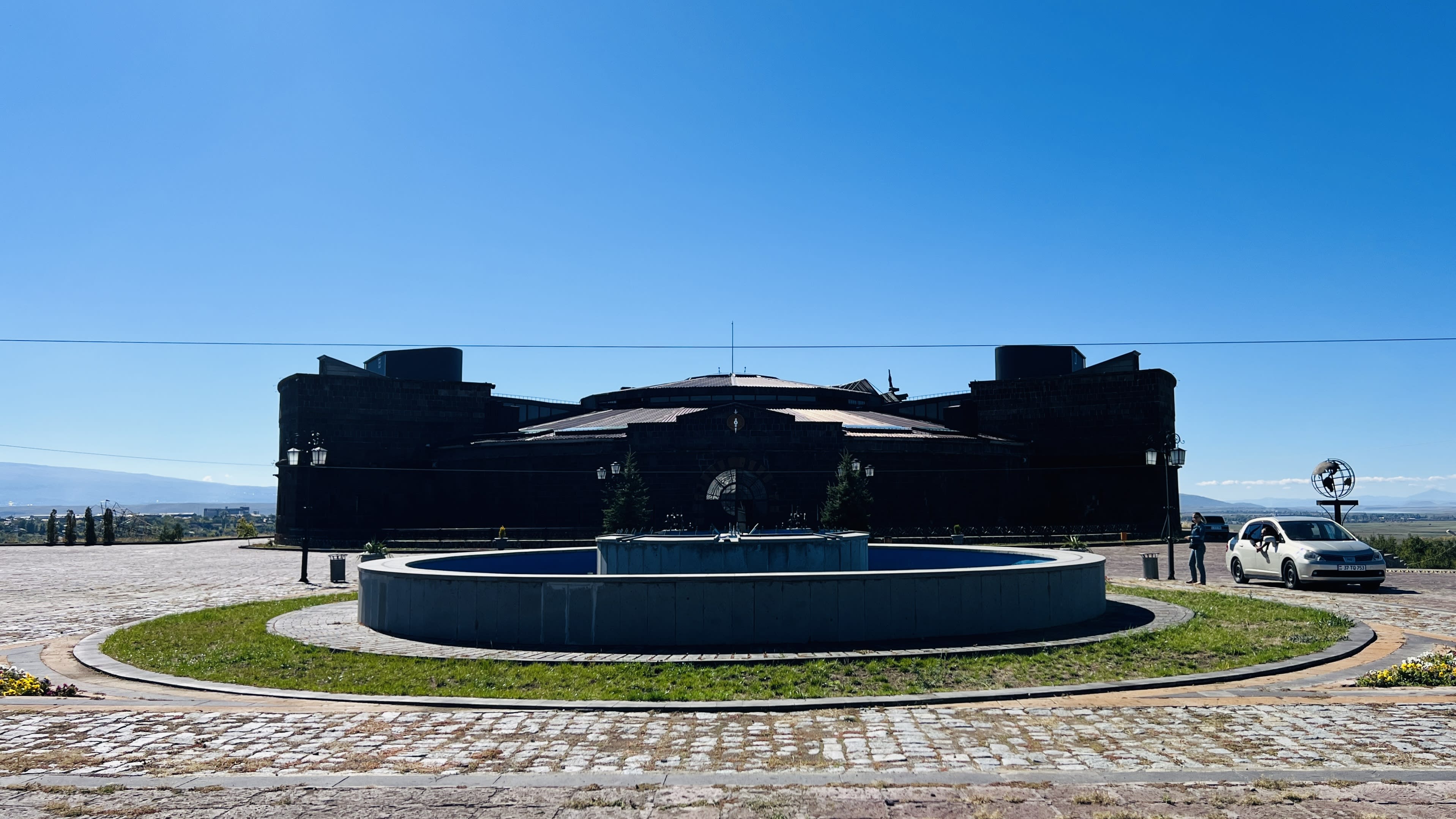 A large stone building with circular and angular features stands against a clear blue sky in Gyumri, Armenia. A white car is parked nearby on a cobblestone path, ready to explore the top gyumri things to do in this charming city.