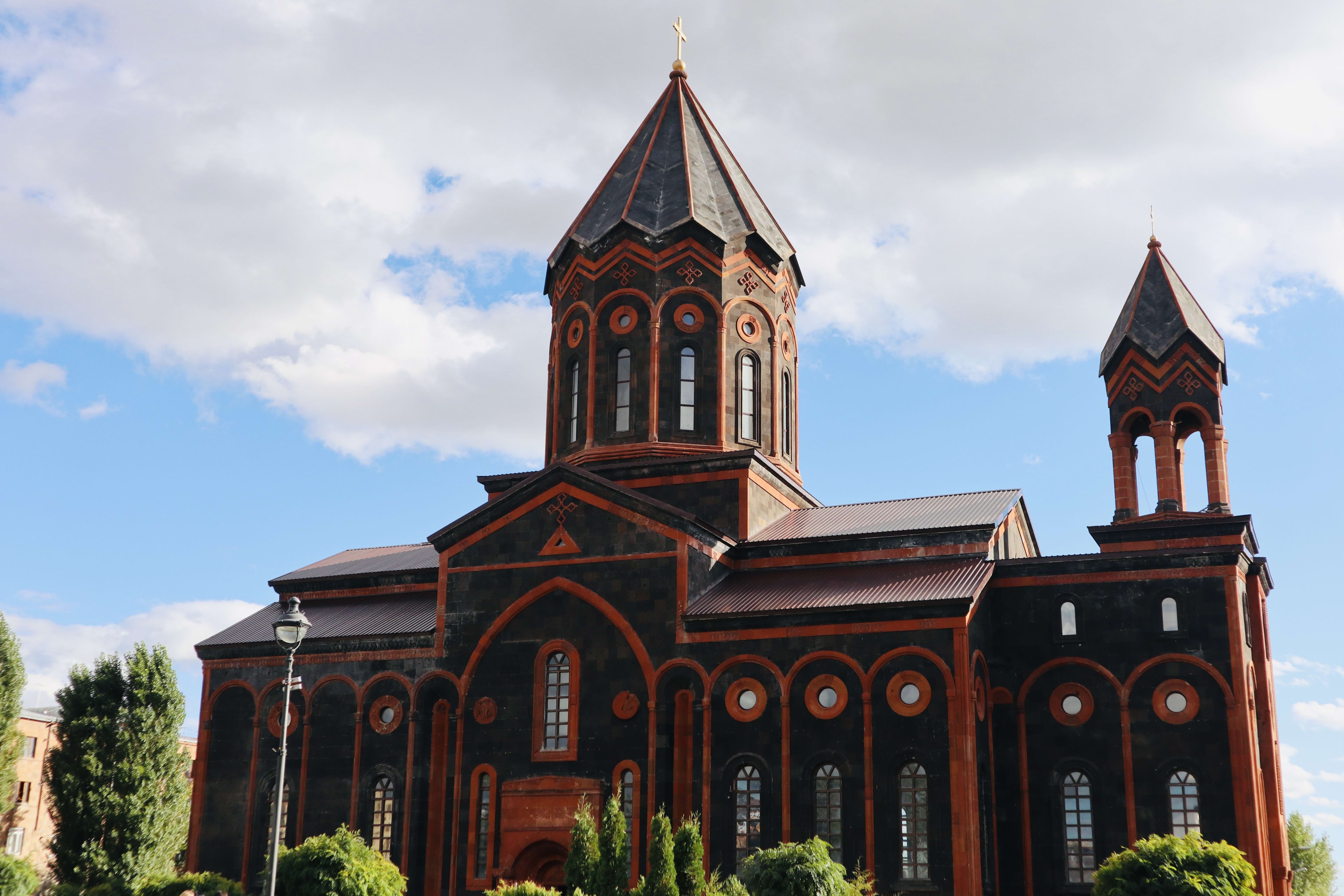 A church in Gyumri with dark stone walls and red detailing stands majestically against a partly cloudy sky in Yerevan. Two towers rise above the structure, while trees grace the foreground, inviting both cultural explorers and those following a Gay travel guide to experience its unique allure. For LGBTQ+ travelers, Gay Yerevan offers a blend of historical sights and inclusive experiences that enrich their journey.