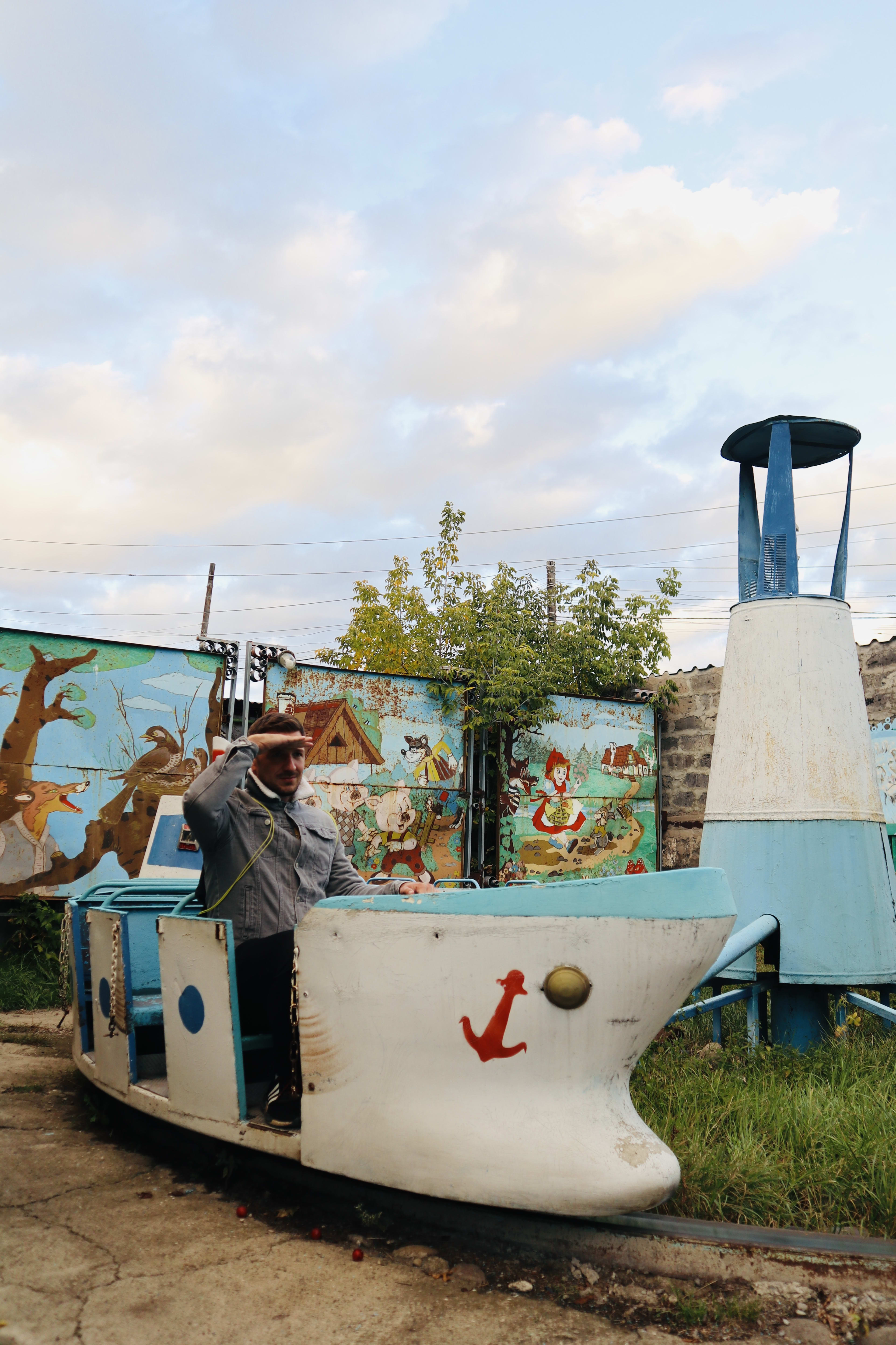 In Gyumri, one of the unique highlights among 'gyumri things to do' is to sit in a small, stationary boat-shaped ride in an outdoor setting. As you salute from the boat featuring a painted anchor, you can admire the mural and blue and white structure behind, creating a memorable experience in Armenia's charming city.