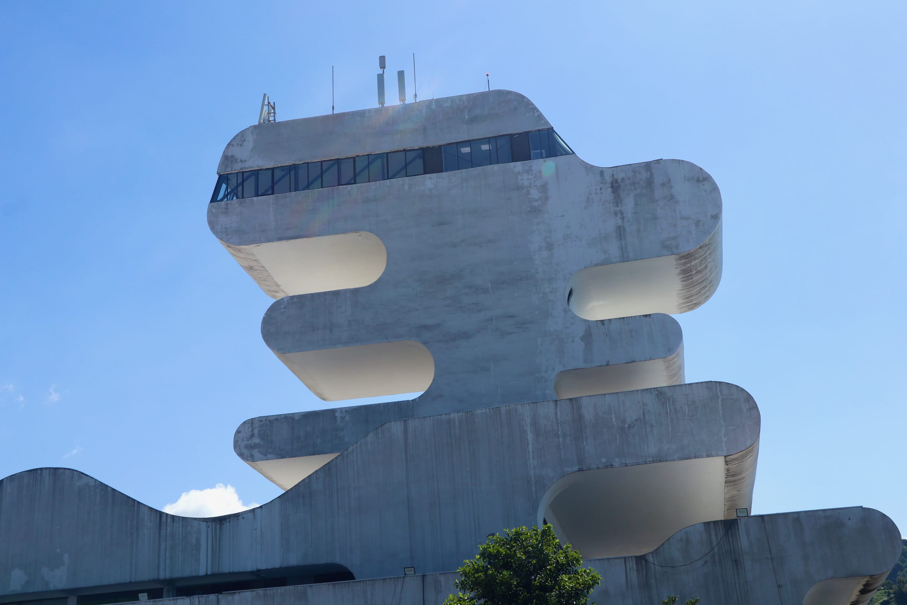 A modern building in Batumi, with a unique, stacked design featuring curved, horizontal layers, set against a clear blue sky. The top layer has a windowed section, and a small tree is visible at the bottom of the image, creating an inviting atmosphere for exploring LGBTQ+ hotspots nearby.