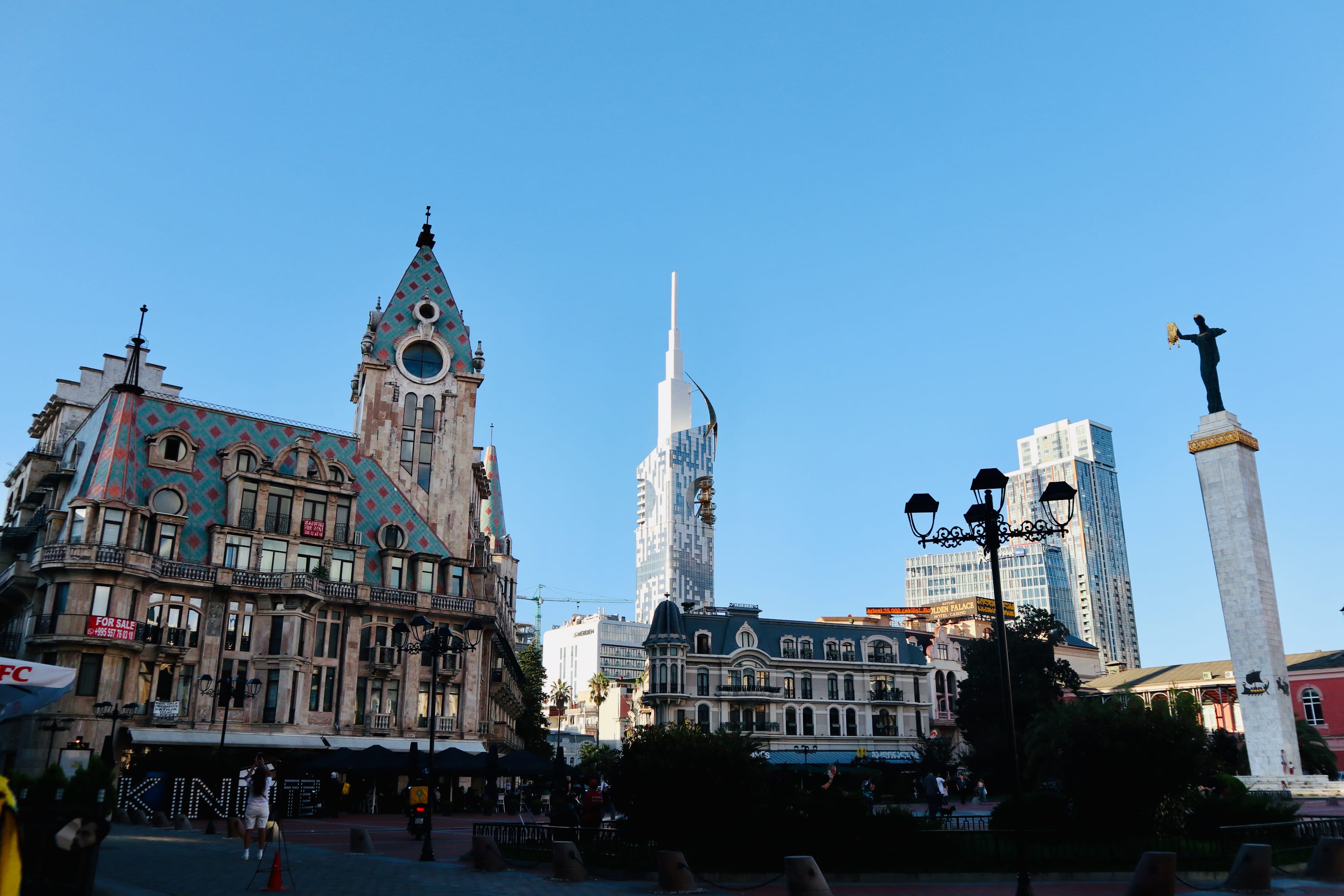 In the vibrant cityscape of Batumi, a decorated historic building with a pointed roof graces the left, contrasting with a modern skyscraper at the center. On the right, a statue atop a tall column completes this picturesque scene under a clear blue sky—a perfect starting point for any gay guide exploring local LGBTQ+ hotspots.