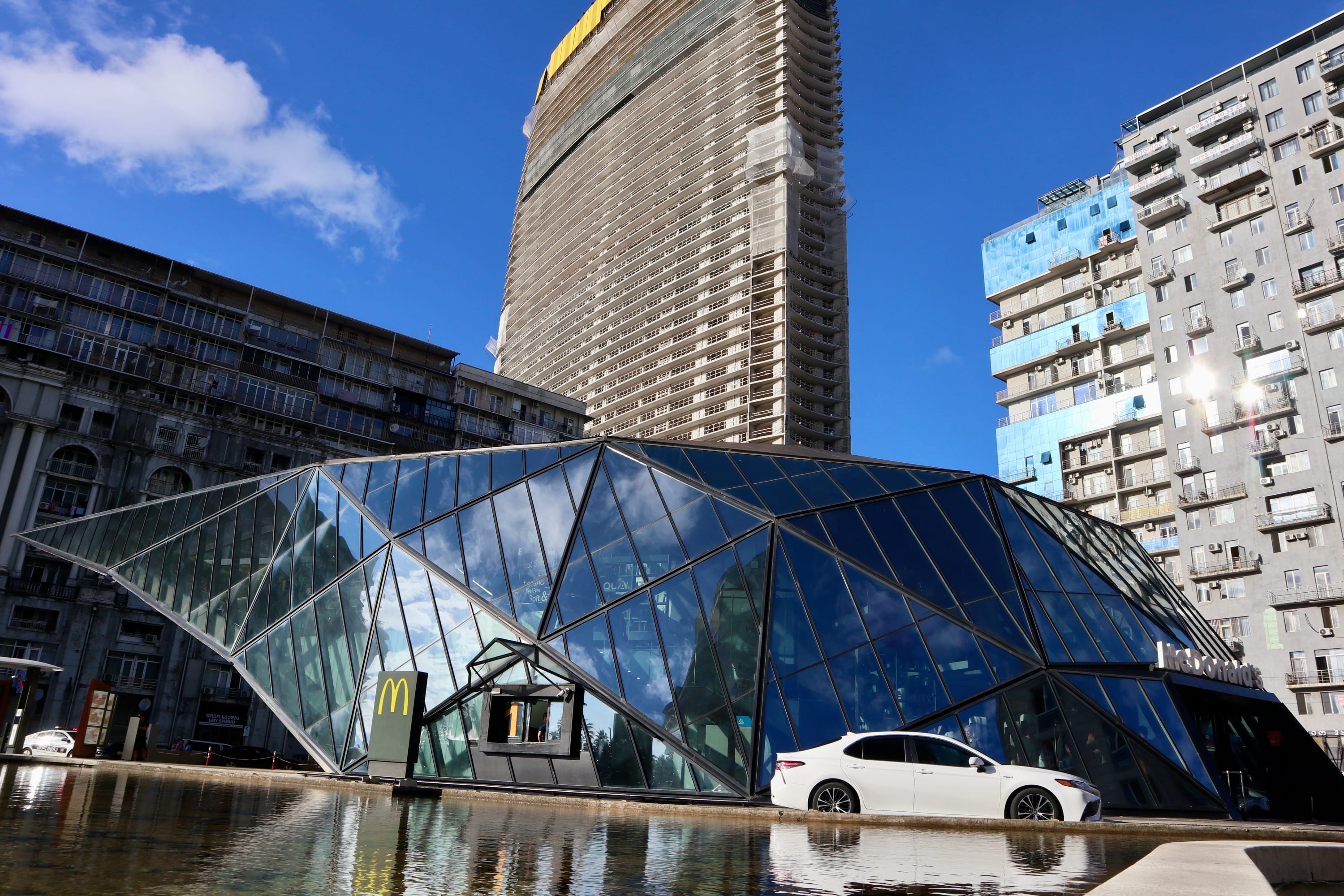 A white car is parked beside a modern, glass-paneled McDonald's building with a unique geometric design in an area noted for its vibrant LGBTQ+ hotspots. Amidst towering constructions, the clear blue sky sets the scene in this colorful urban landscape.