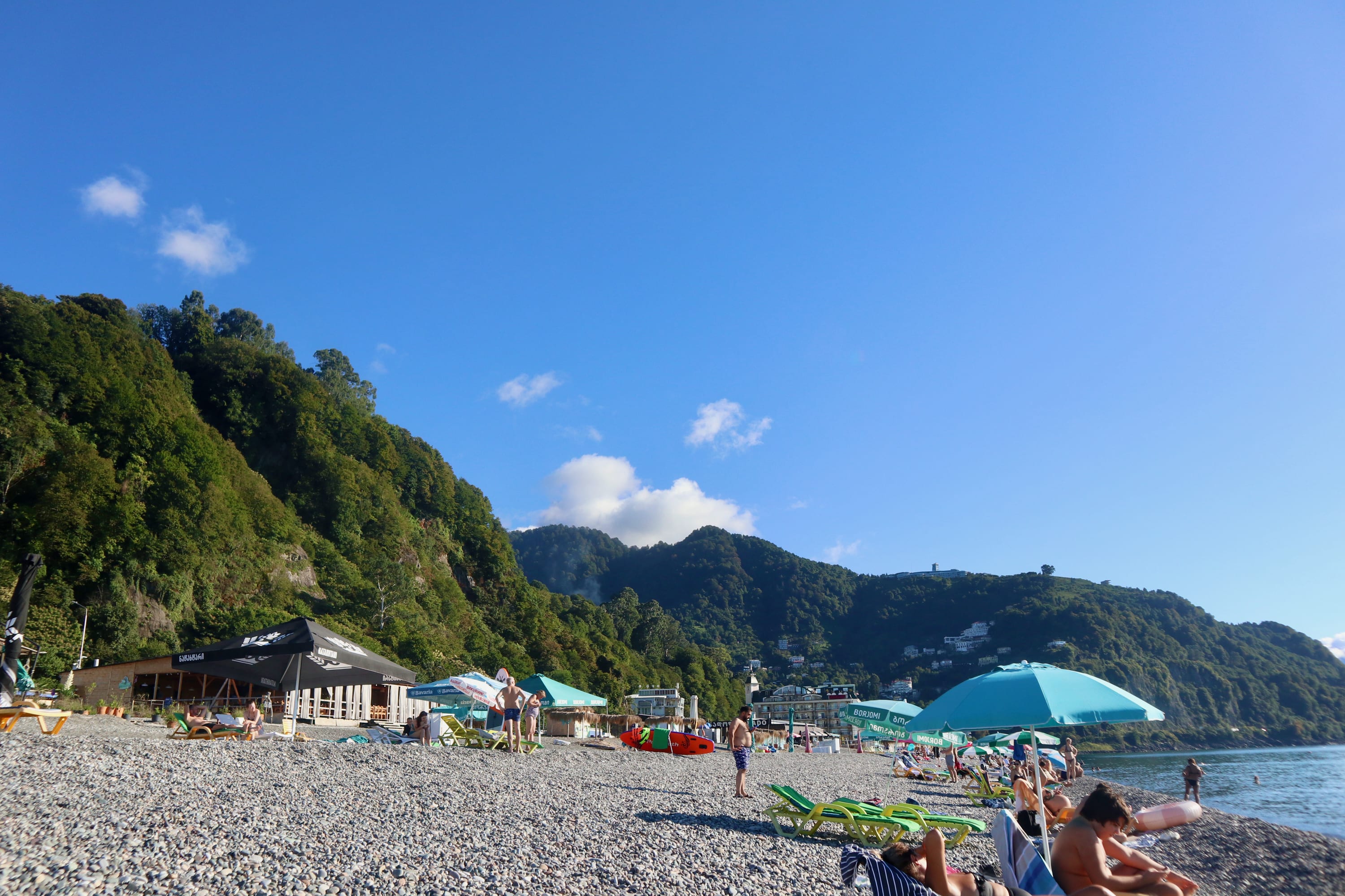 A pebble beach in Batumi, where people relax under blue umbrellas. The backdrop features lush green hills and a clear blue sky, offering a picturesque setting often highlighted in many gay guides as one of the must-visit LGBTQ+ hotspots. A small building and beach chairs dot the shore.
