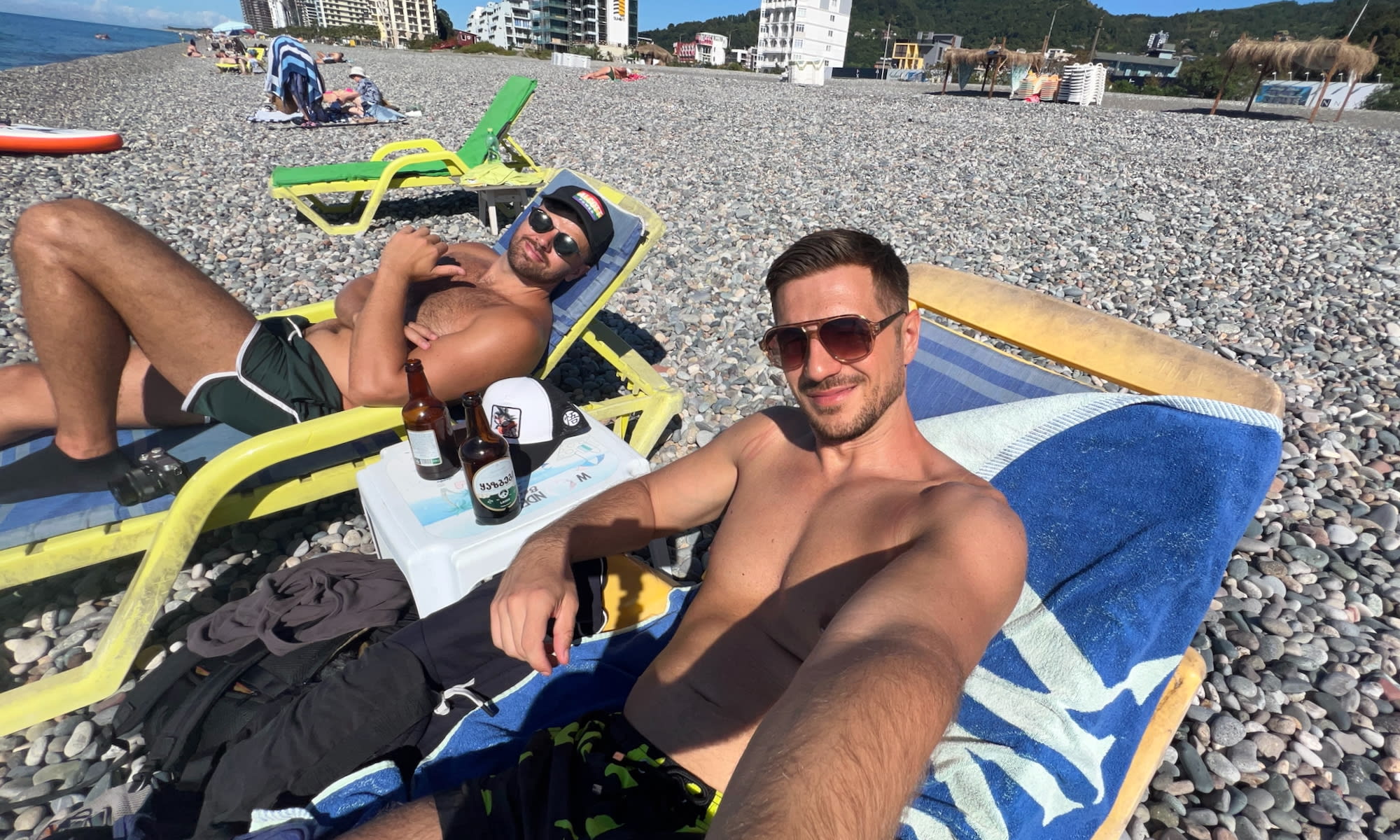 Two men are sunbathing on a pebble beach in Batumi, relaxing on lounge chairs under a clear blue sky. They are shirtless, wearing sunglasses, and have beverages on a small table between them. Known for its LGBTQ+ hotspots, the city offers vibrant scenes amidst buildings and hillside views.
