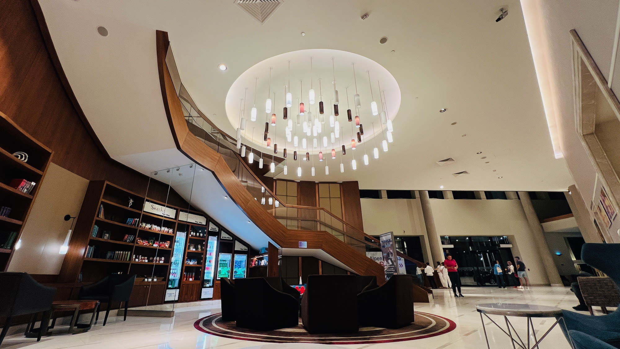 A modern hotel lobby in vibrant Batumi features a large, elegant chandelier. Below, a grand wooden staircase descends amidst dark lounge chairs around a circular carpet, bookshelves lining the walls. In this lively setting that's part of the city's LGBTQ+ hotspots, people gather, adding to its unique charm.