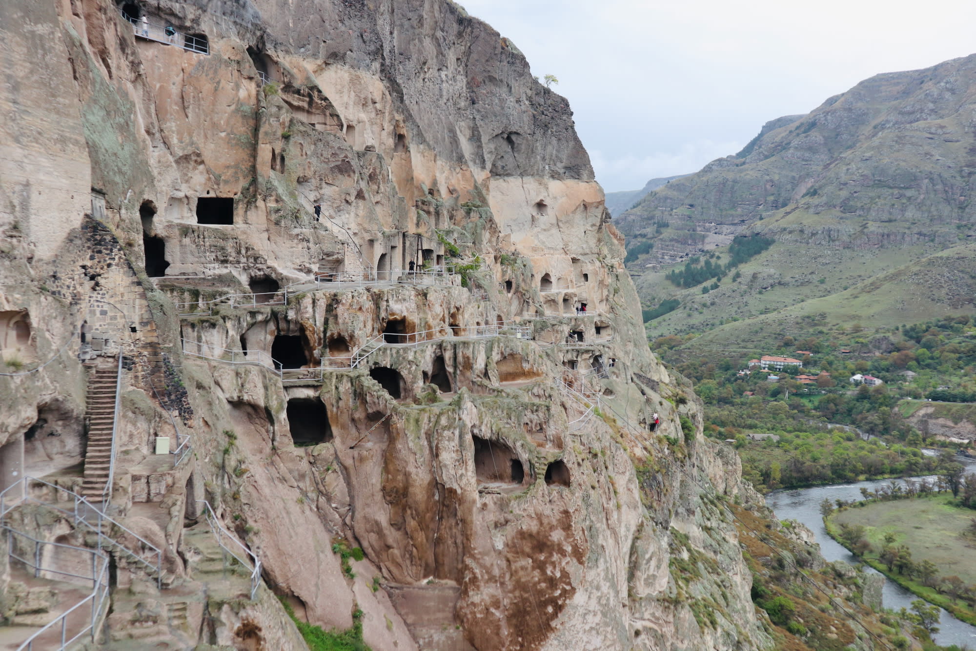 Nestled near Akhaltsikhe, this must-see ancient cave-city boasts multiple openings and pathways carved into the rock. A lush green valley and winding river stretch elegantly under a cloudy sky, showcasing one of the region's cultural gems.