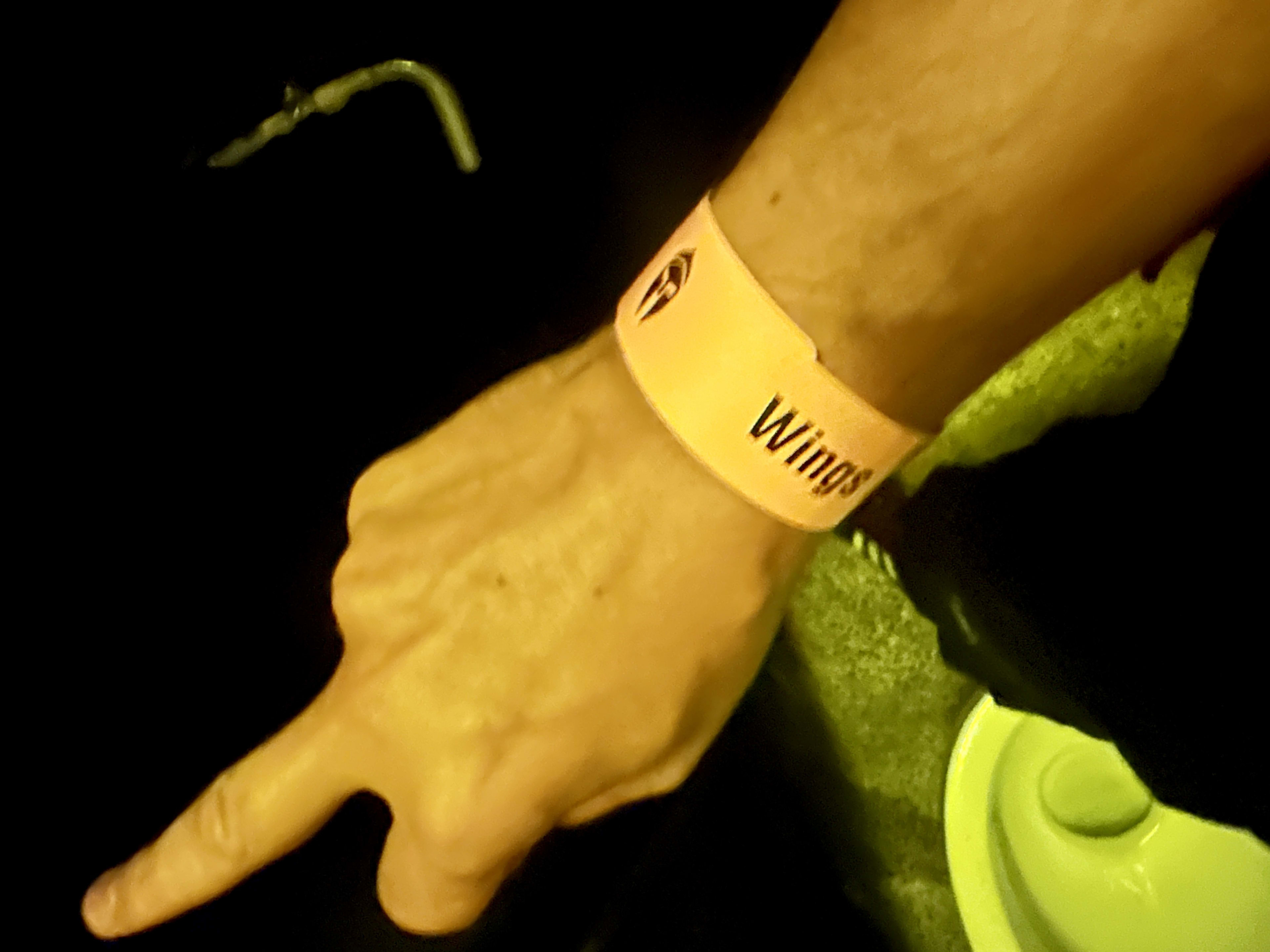 A person's left hand is pointing downward in a dimly lit setting, reminiscent of Tbilisi nightlife, perhaps even the famed Bassiani club. They are wearing a yellow wristband with the text 'Wings' printed on it. A small part of a white object, possibly a cup or bowl, is visible on the right side of the image.
