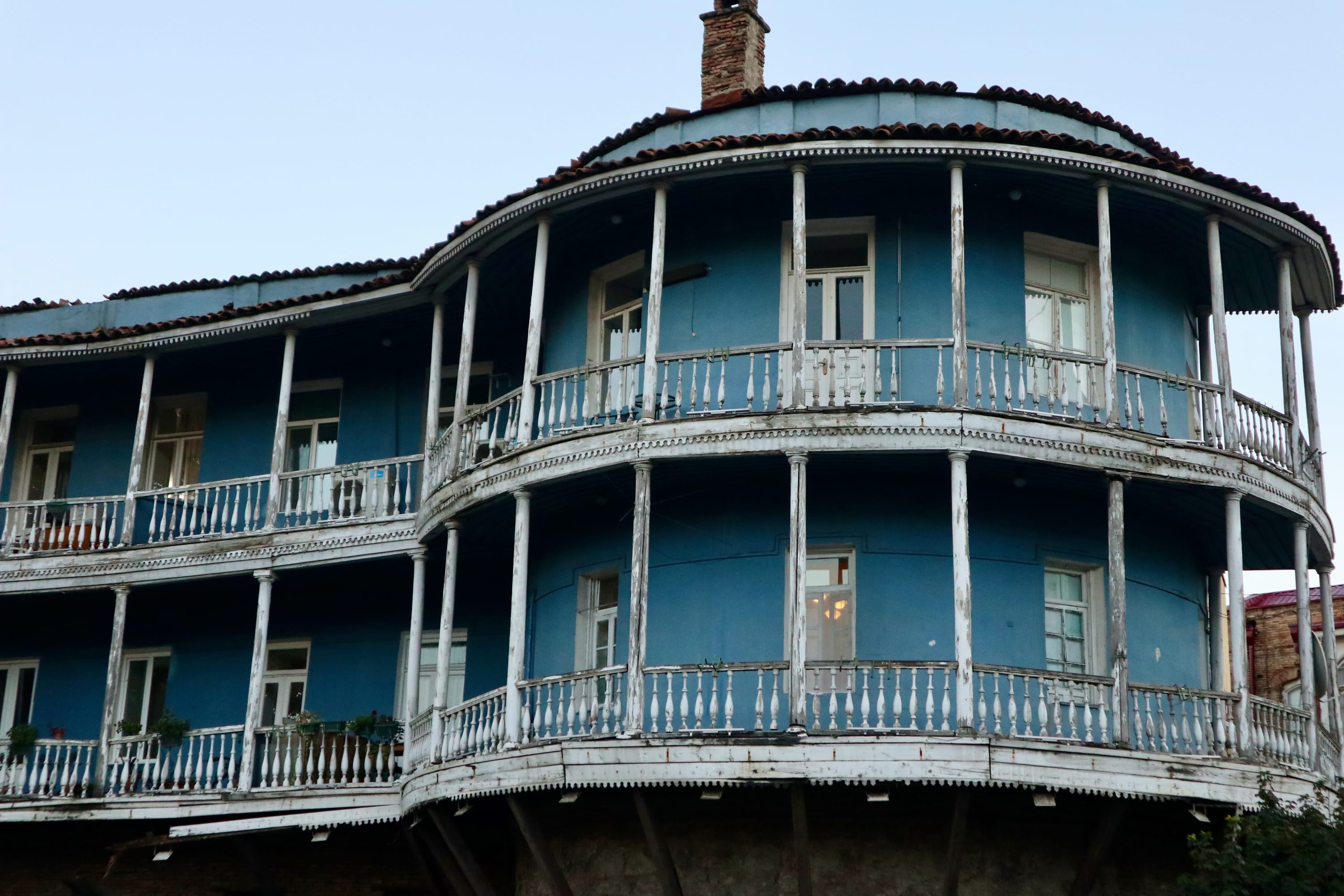 A blue three-story building in Georgia boasts curved balconies and a weathered white railing, with multiple windows offering unique travel experiences. A charming chimney completes its inviting look, making one wonder: is Georgia worth visiting?