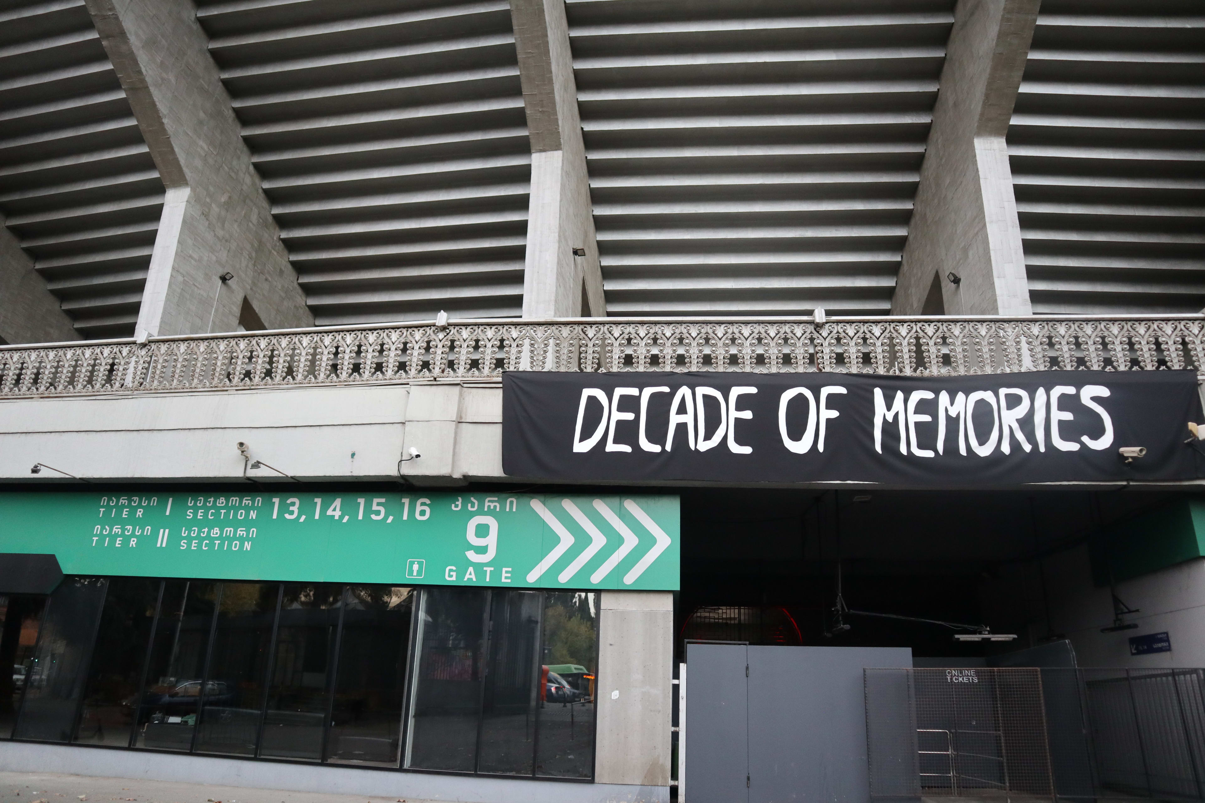 Exterior view of the Bassiani nightclub entrance at Dinamo Arena in Tbilisi, featuring a large black banner with white text reading 'DECADE OF MEMORIES' hung above Gate 9. The green and white signage below indicates sections and tiers in both Georgian and English.