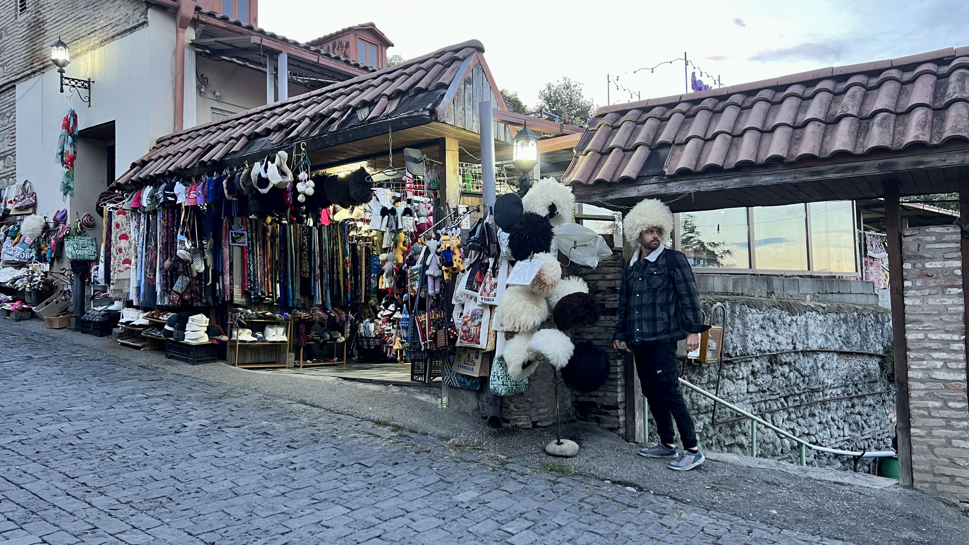A person stands by a street stall displaying hats, scarves, and other items on a sloped cobblestone street in the romantic town of Sighnaghi, Georgia.