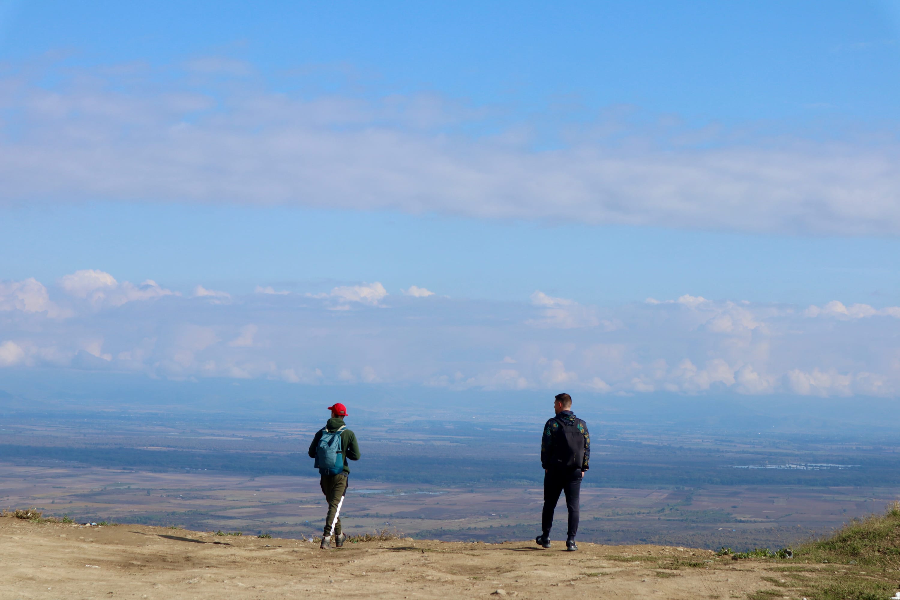 Two people with backpacks stand on a dirt path in Sighnaghi, the City of Love, looking out over a vast Georgian landscape with a clear blue sky and distant clouds. If you're wondering about things to do in Sighnaghi, Georgia, consider exploring the charming cobblestone streets lined with quaint houses and stunning views of the Alazani Valley. Visit the historic Bodbe Monastery for its spiritual ambiance and beautiful gardens or take a romantic walk on the ancient city walls for panoramic vistas. Don't forget to sample local wines from renowned vineyards in this picturesque region.
